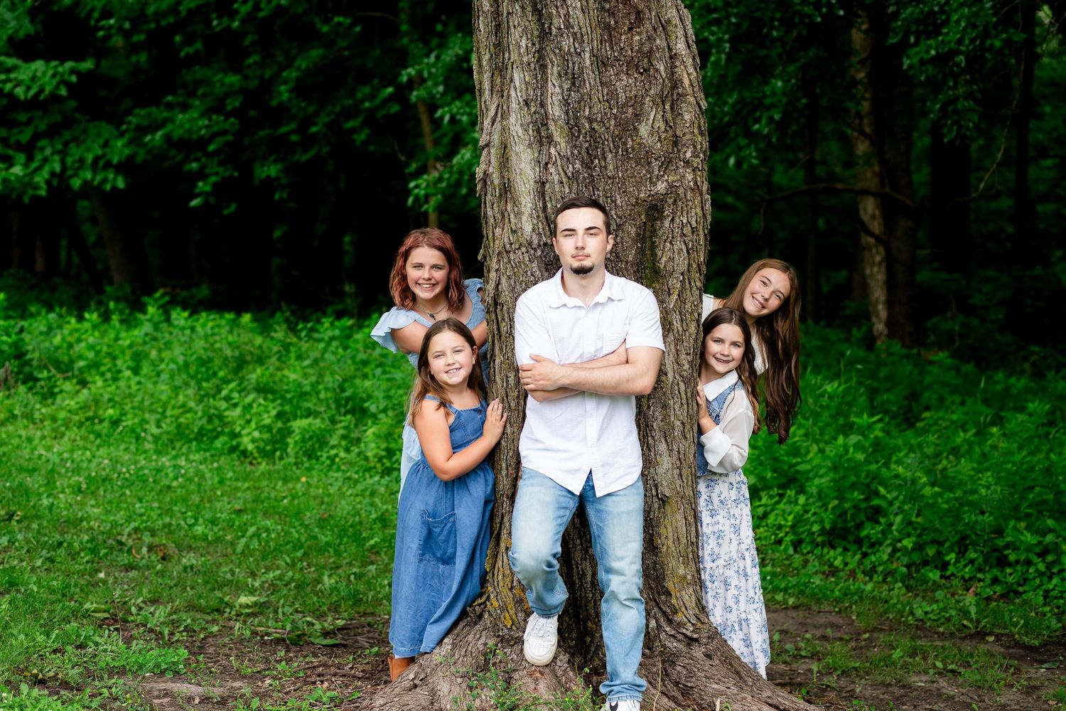 Family of five posing around a large tree trunk in a lush green forest setting.