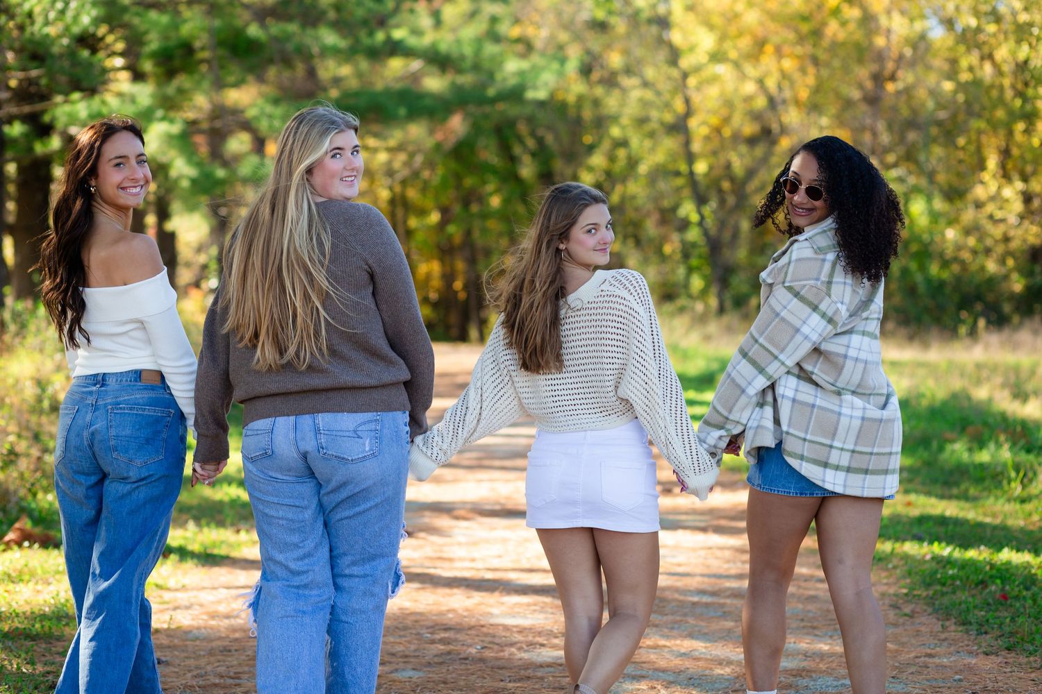 High school senior girls walking together on a sunlit nature path at Pinicon Ridge surrounded by green trees and grass on a warm summer day.