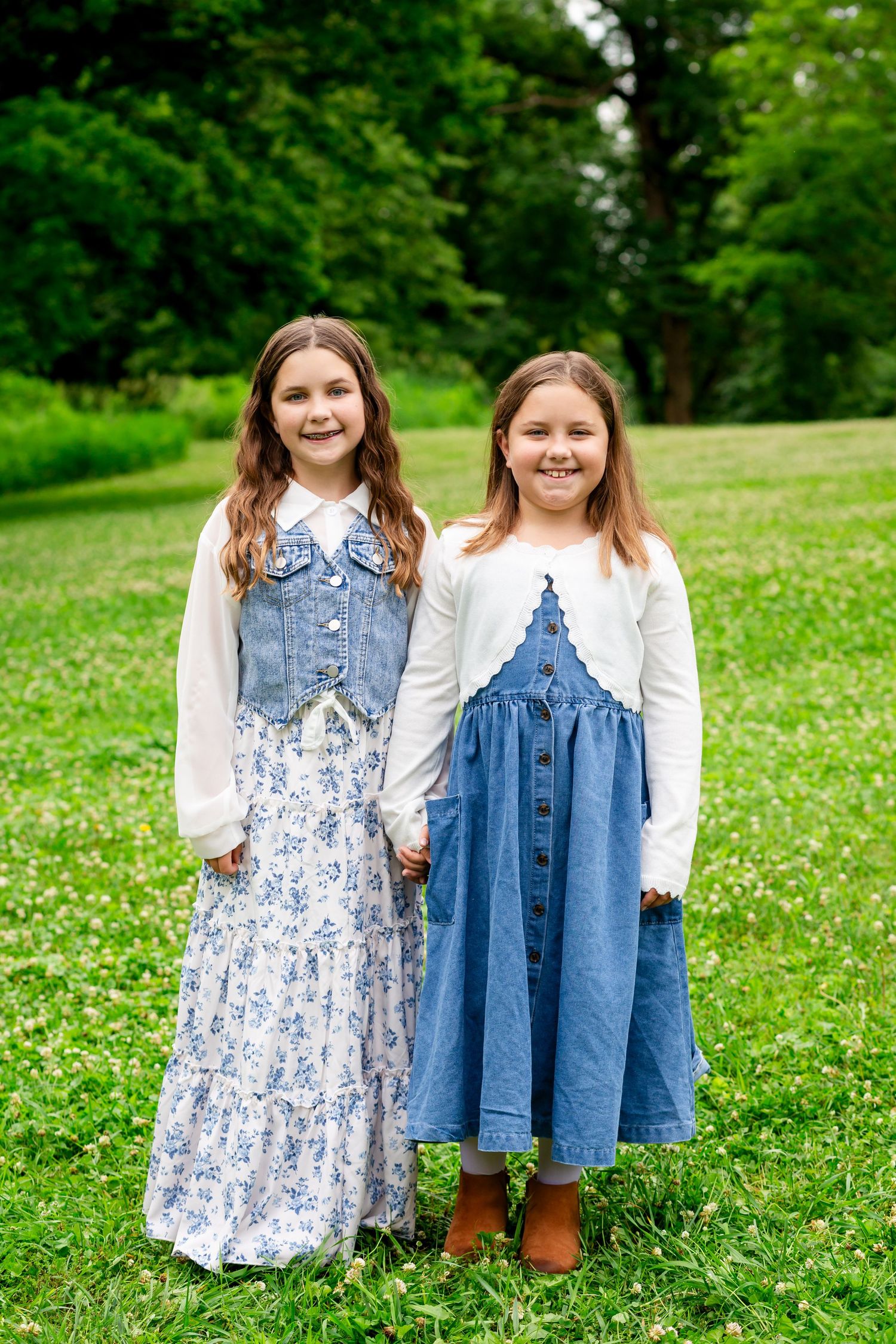 Two young girls in denim skirts holding hands and smiling on a green grassy field.