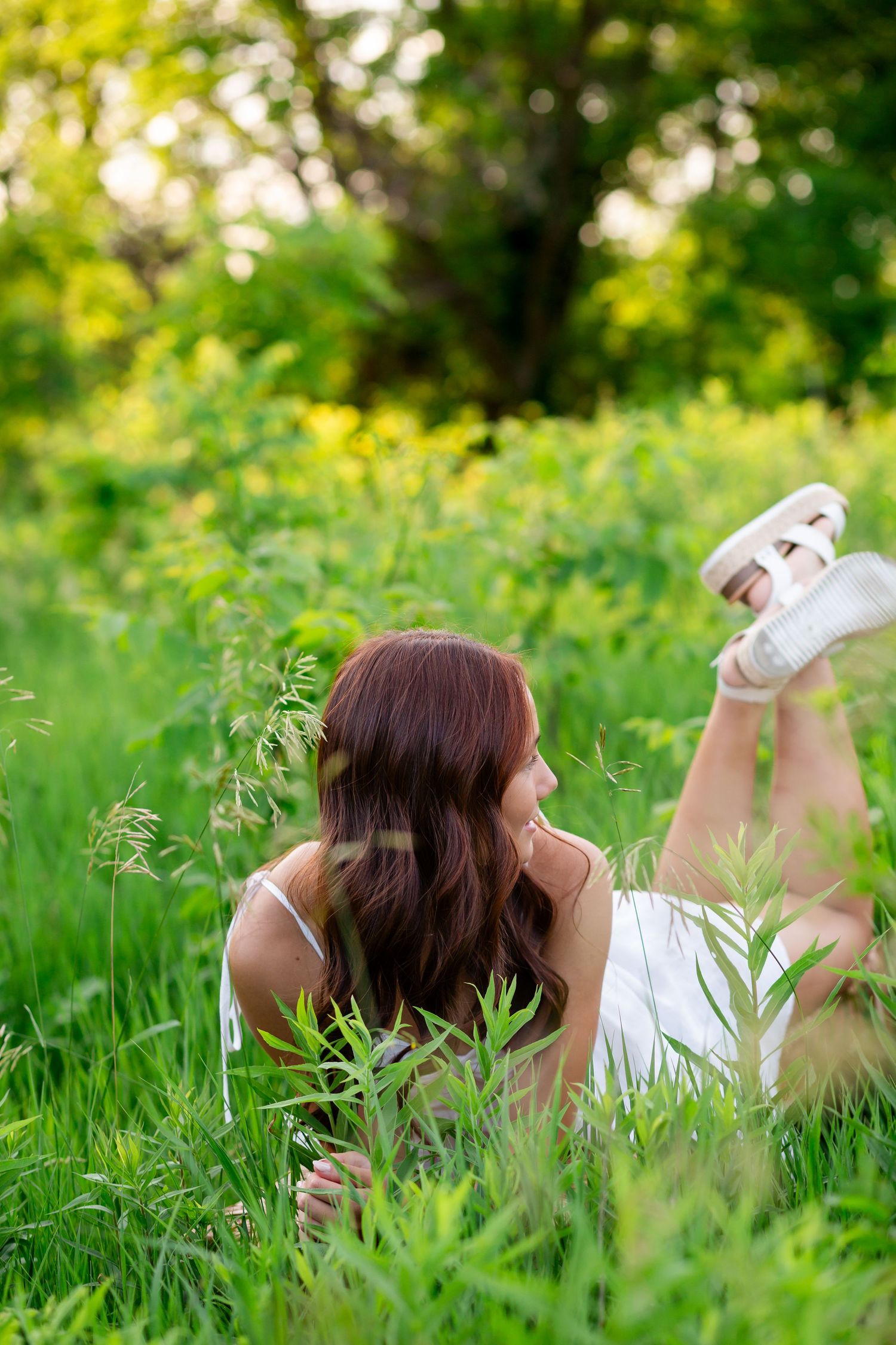Young woman with long brown hair lying in lush green meadow grass wearing white dress and sandals.