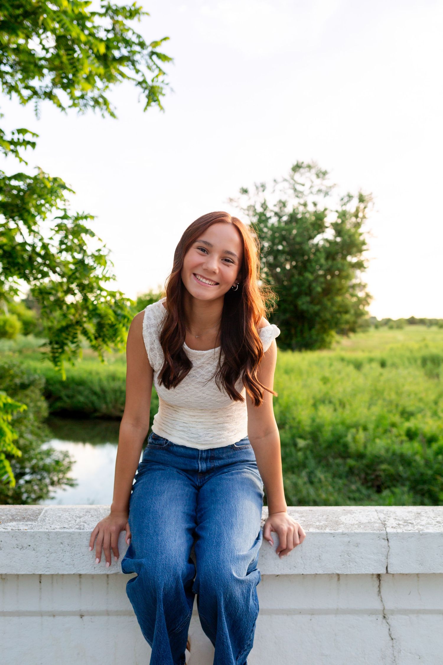 Smiling teenage girl with long brown hair sits on white stone ledge in jeans and white top outdoors.