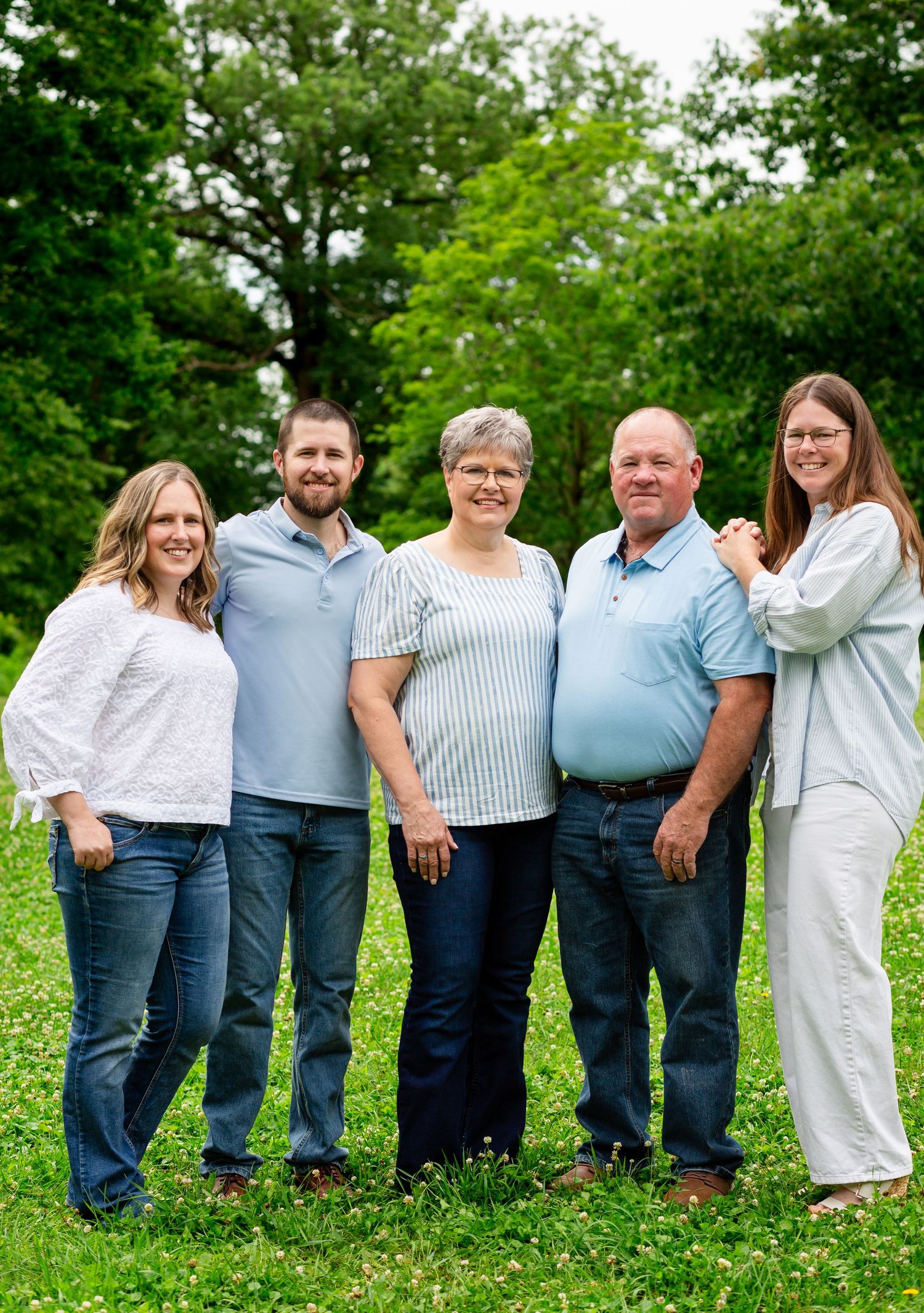 Family of five smiling together outdoors on a green grassy hill surrounded by trees.