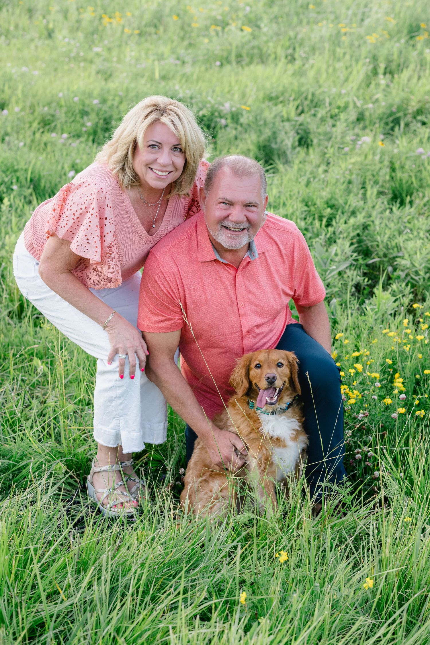 Smiling couple in matching coral tops posing with golden retriever dog in lush green meadow with yellow wildflowers.