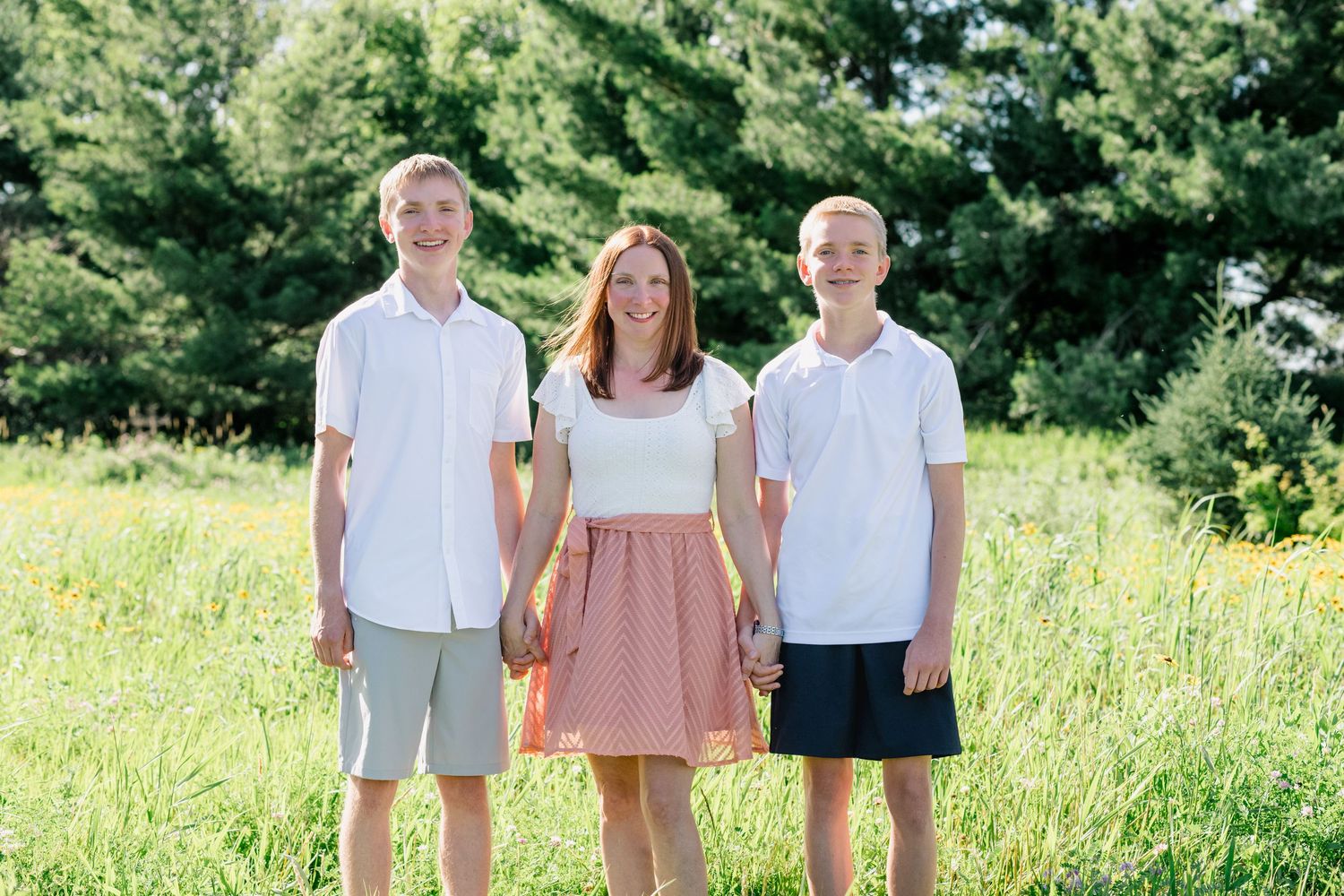 Three smiling teens pose outdoors in a sunny green field, two boys in white polos and a girl in a pink skirt.