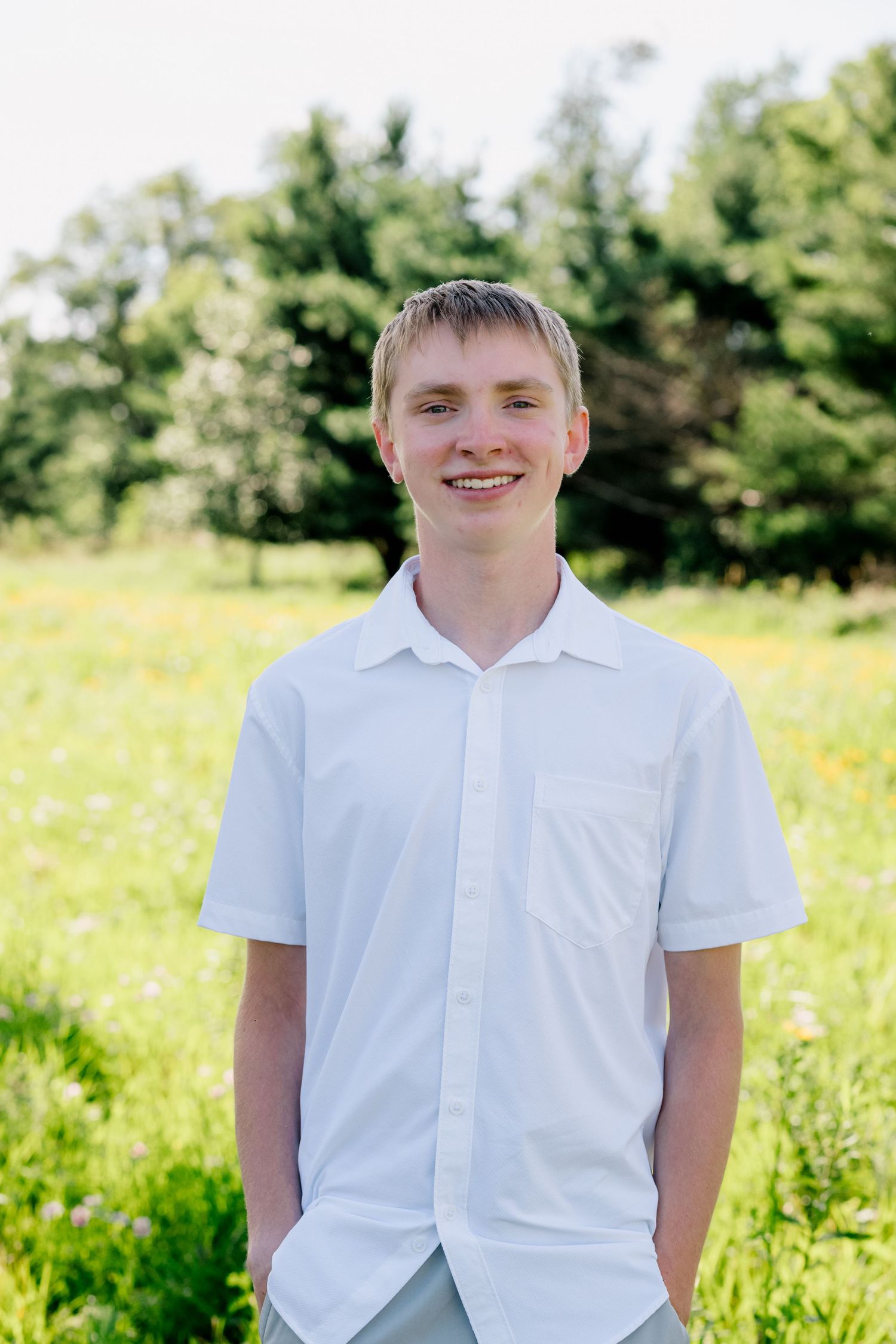 Smiling teenage boy in white button-up shirt standing in green meadow with trees.