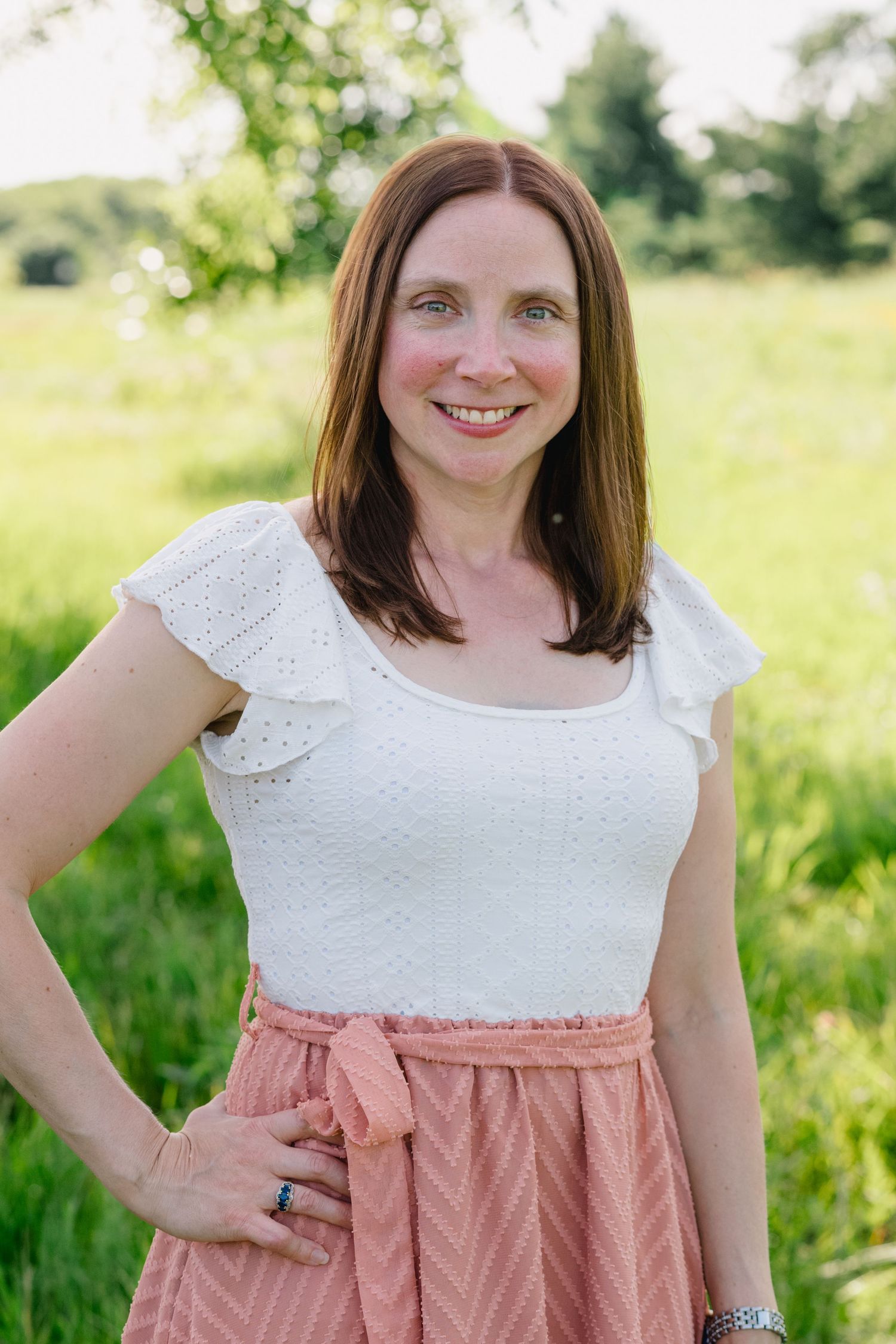 Smiling woman with brown hair in white top and pink skirt posing in sunny meadow.
