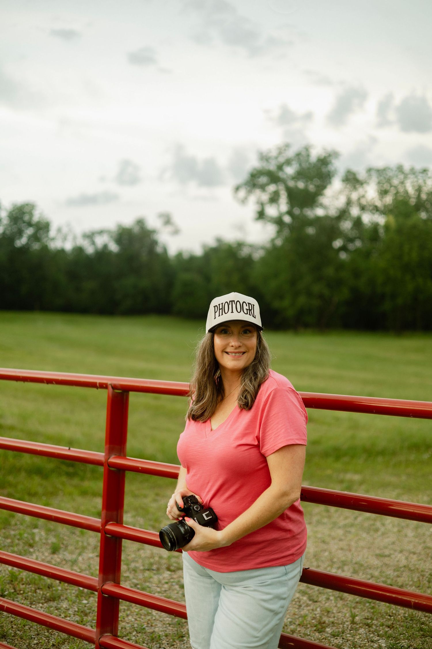 Woman photographer in pink shirt and trucker hat holds camera while leaning on red farm gate in green field.