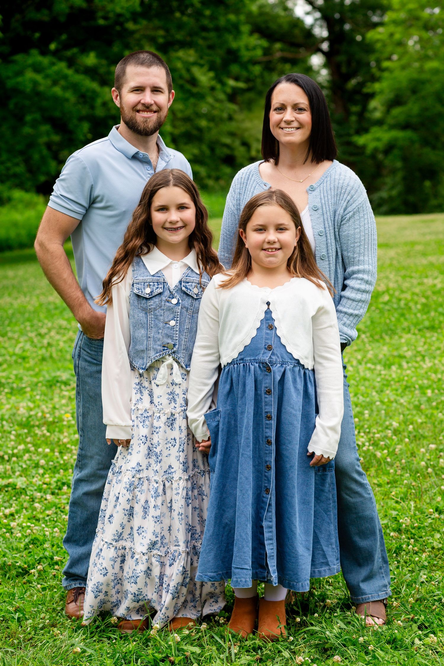 Family of four posing outdoors on green grass, parents standing behind two daughters in denim dresses.