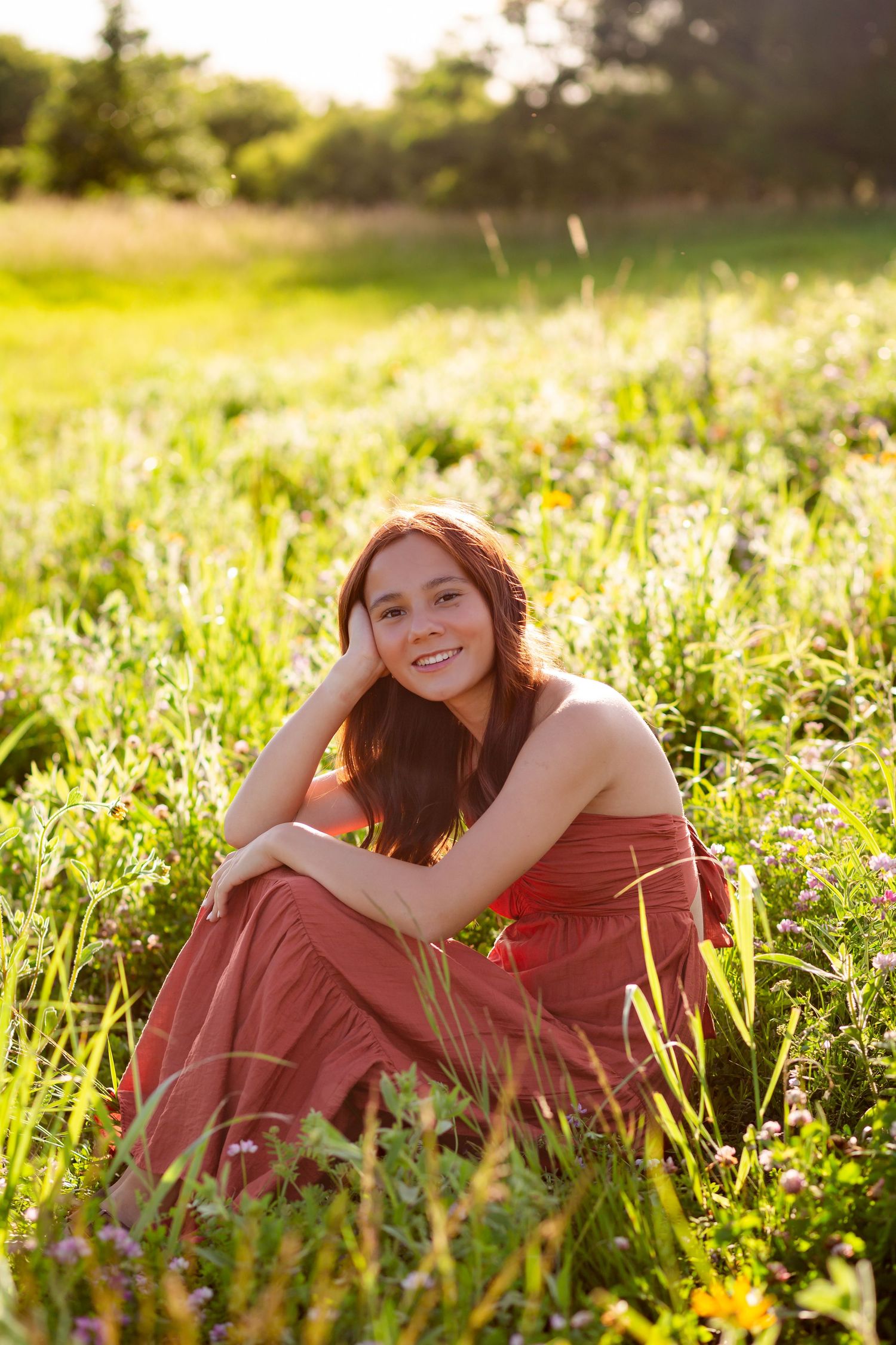 Smiling young woman in red dress sitting in a sunny wildflower meadow, resting her head on her hand.