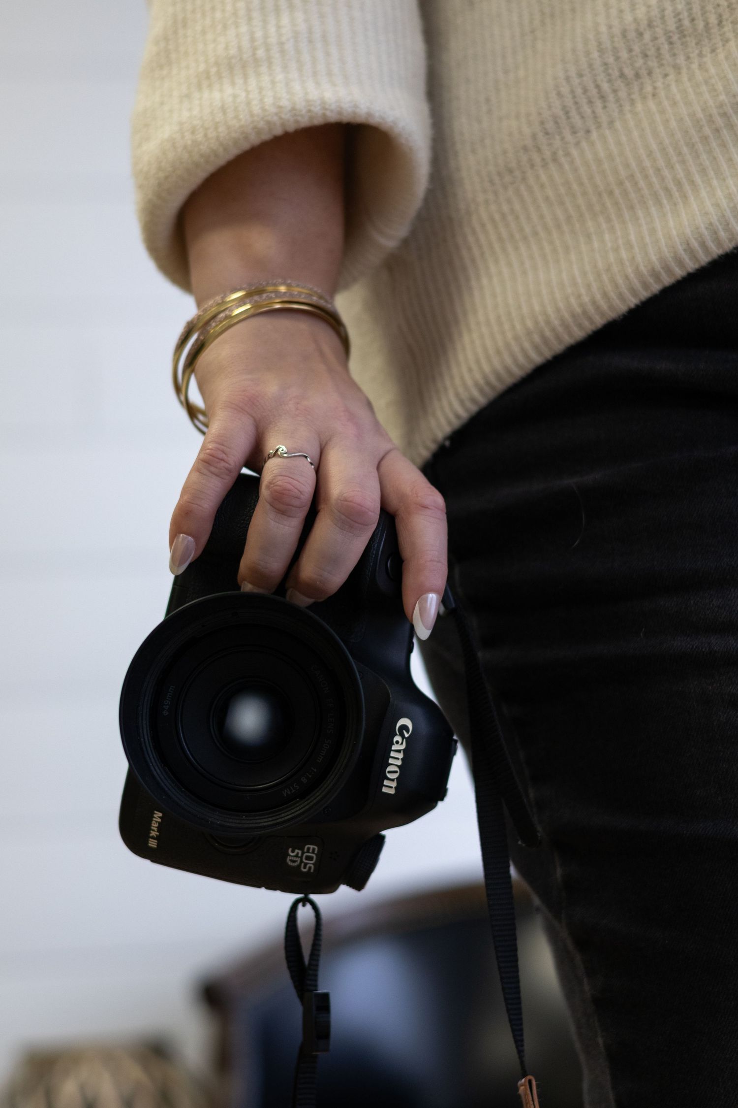 Hand wearing golden jewelry holds a Canon camera against a cream sweater and black pants background.