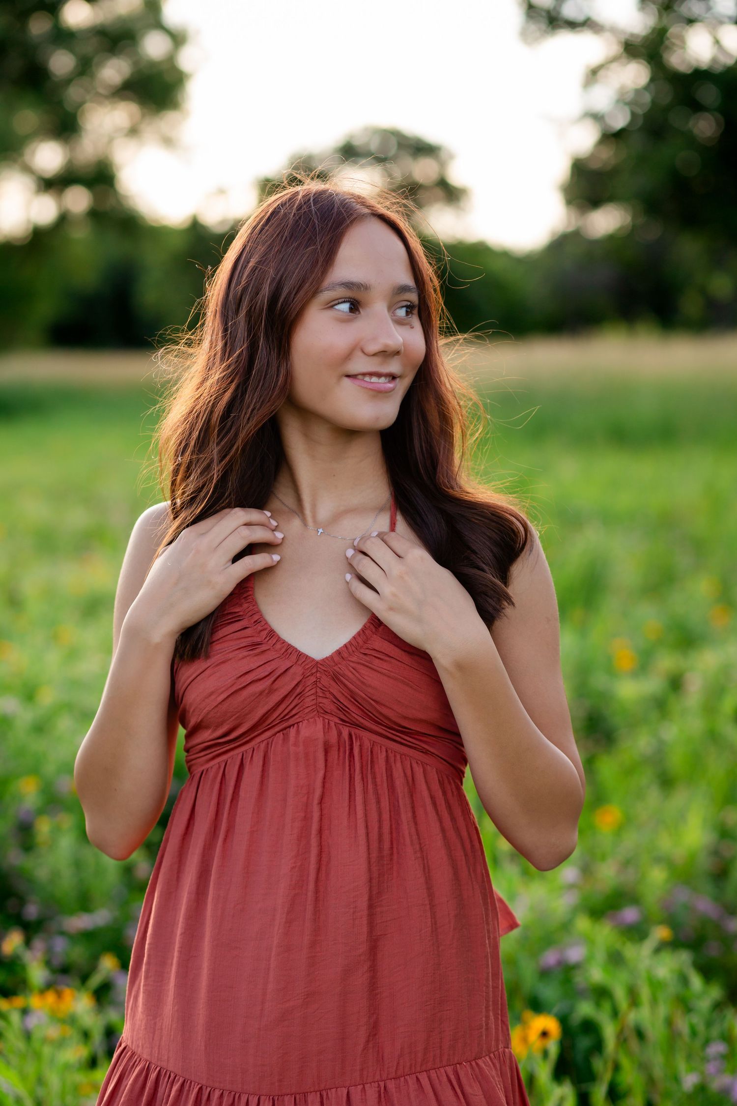 Young woman in rust red dress smiling in a green field at golden hour, hands resting on her chest.