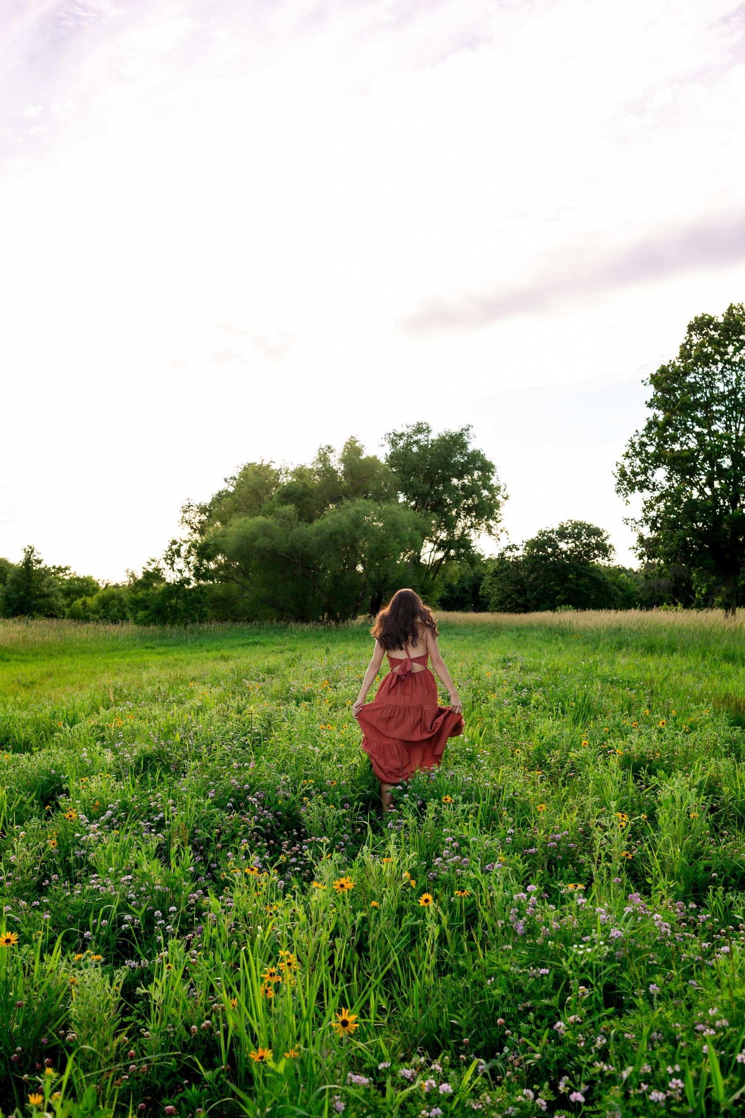 Woman in flowing red dress walks through lush green wildflower meadow at dusk with trees in background.