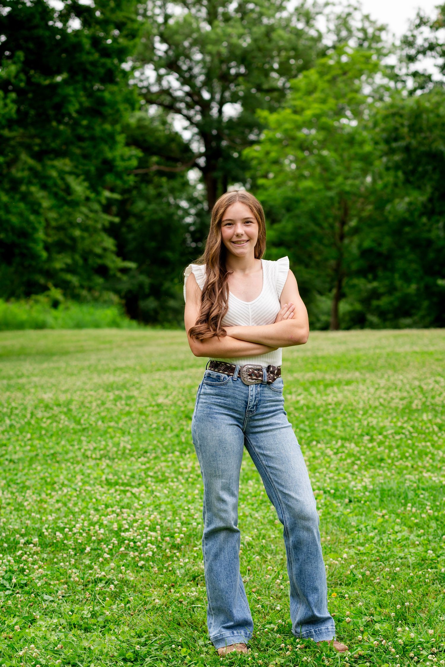 Teenage girl in white top and flared jeans standing confidently with arms crossed in green park.