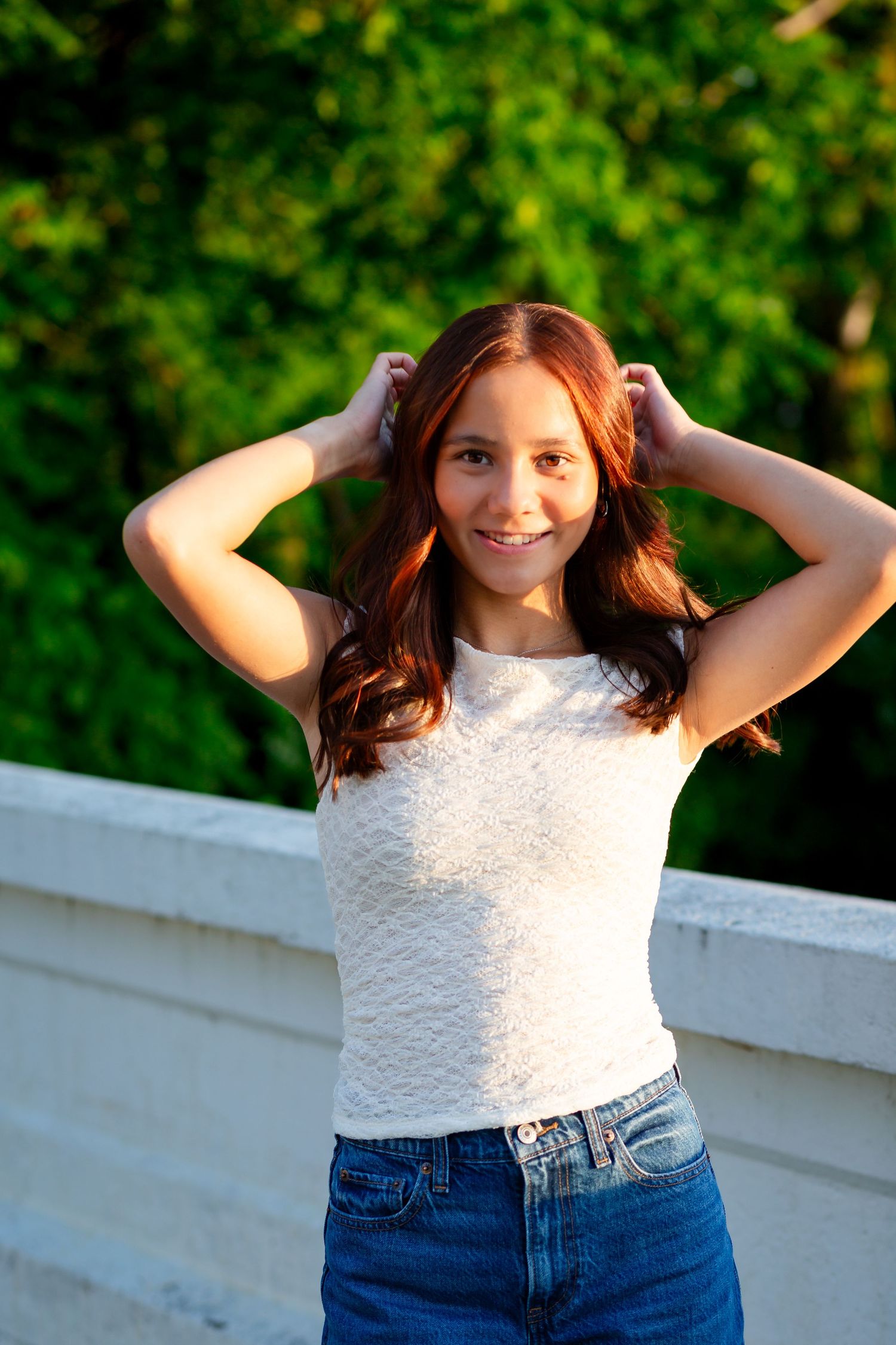 Smiling teenage girl with red wavy hair in a white top and jeans posing outdoors near green trees.