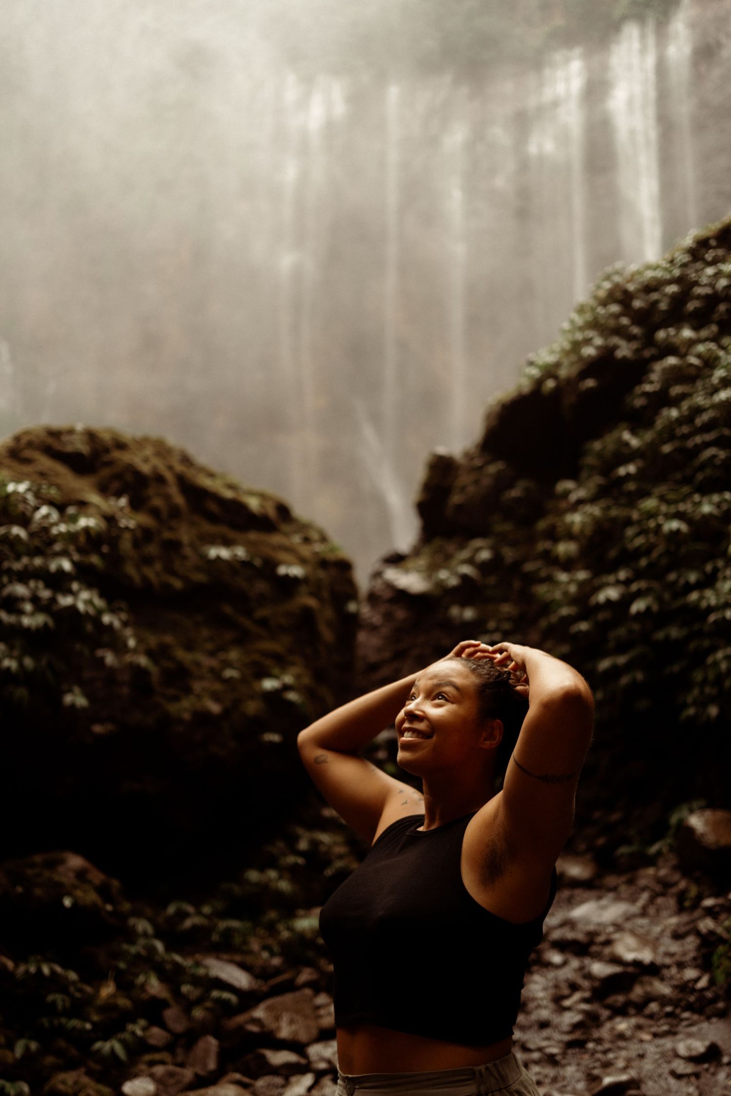Woman in black sports bra smiles with hands in hair before misty tropical waterfall.