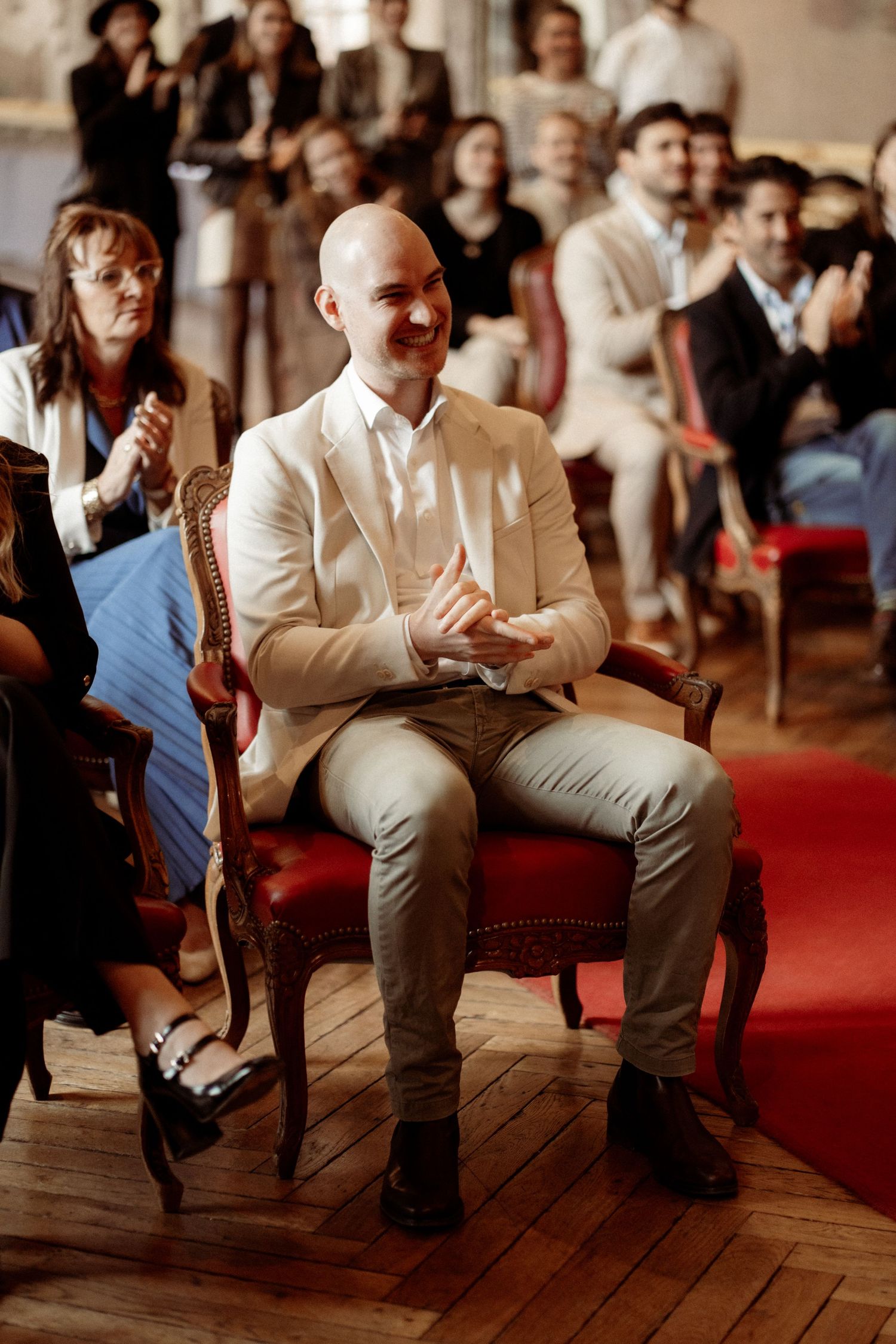 Smiling bald man in beige suit seated in antique chair during wedding ceremony with guests applauding.