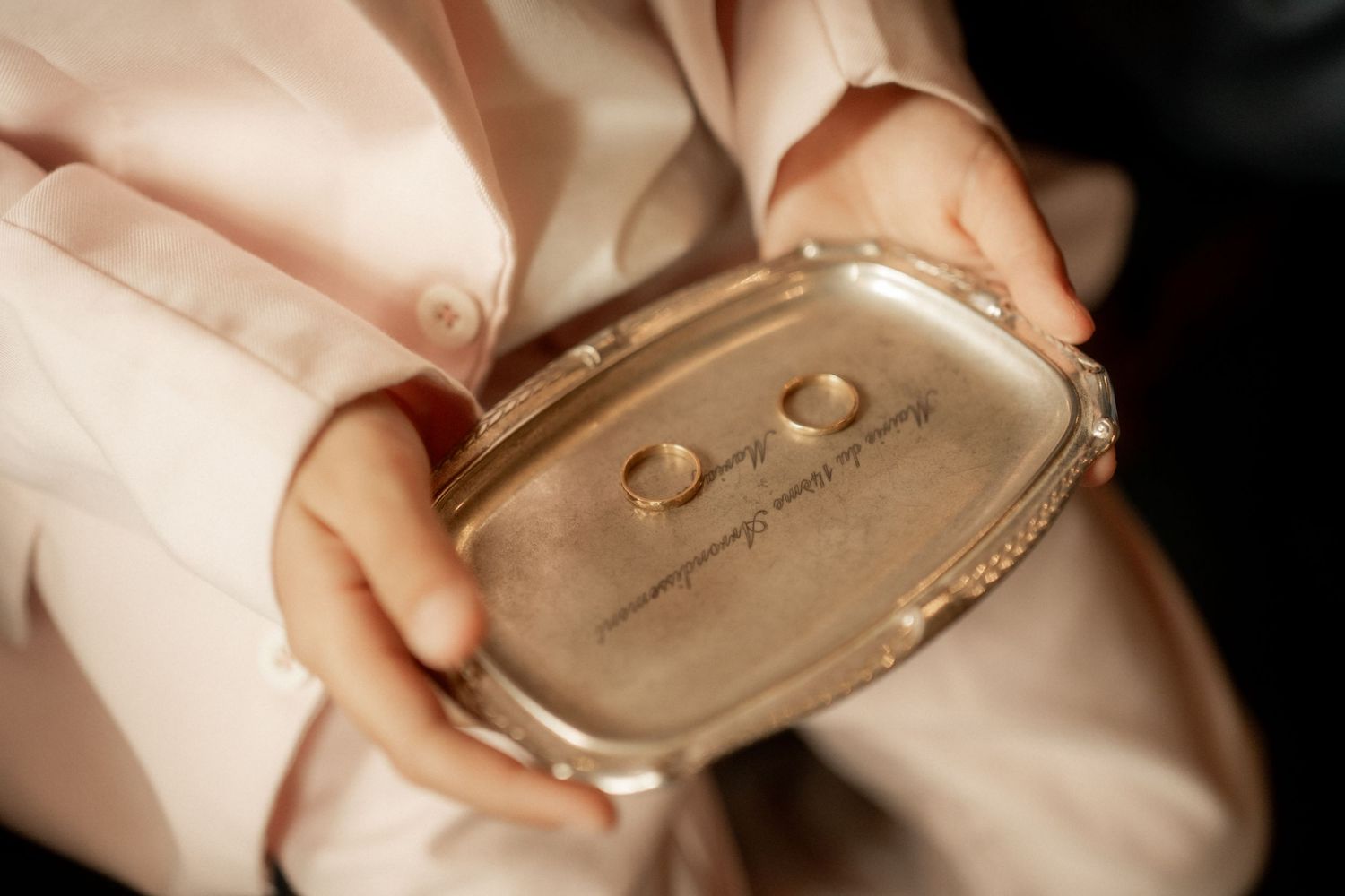 Two gold wedding rings displayed on a silver decorative tray held by hands in a pink satin robe.