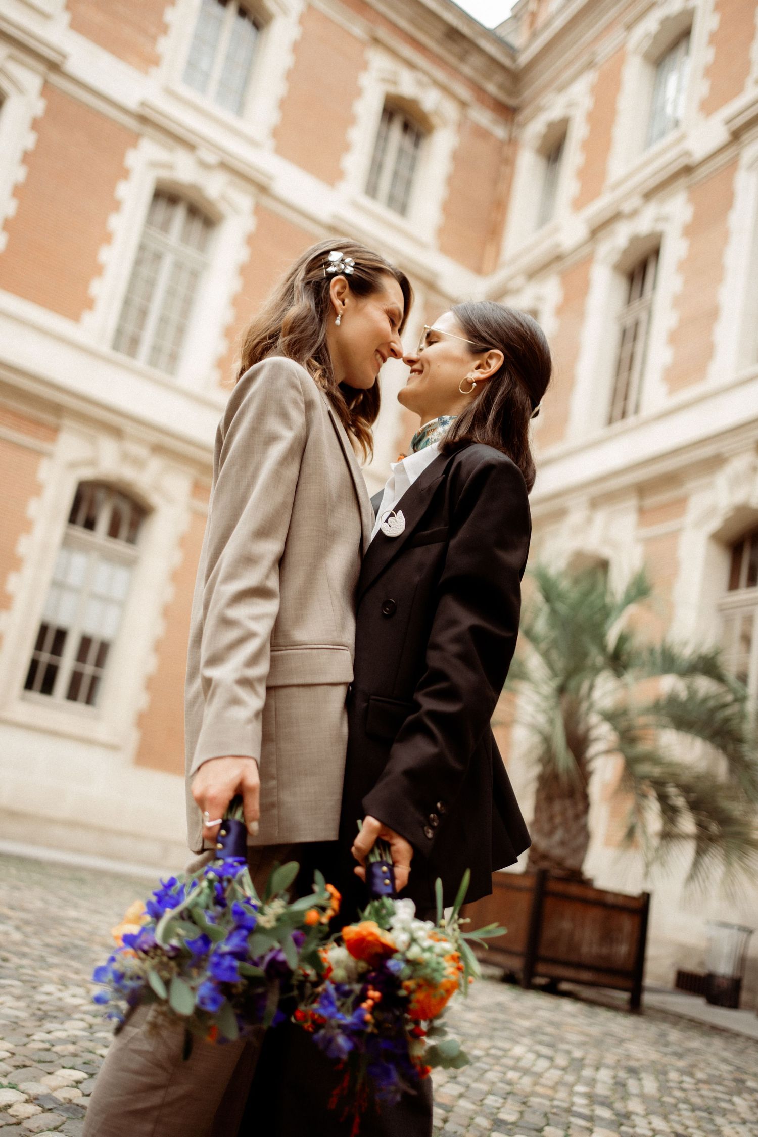 Two brides in suits sharing a romantic moment holding colorful bouquet in a classic European courtyard.