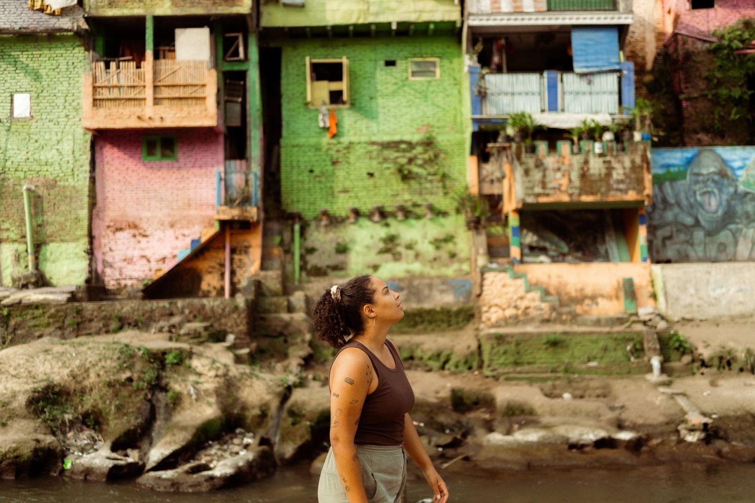 Woman standing by a river with colorful green and pink urban buildings in the background.
