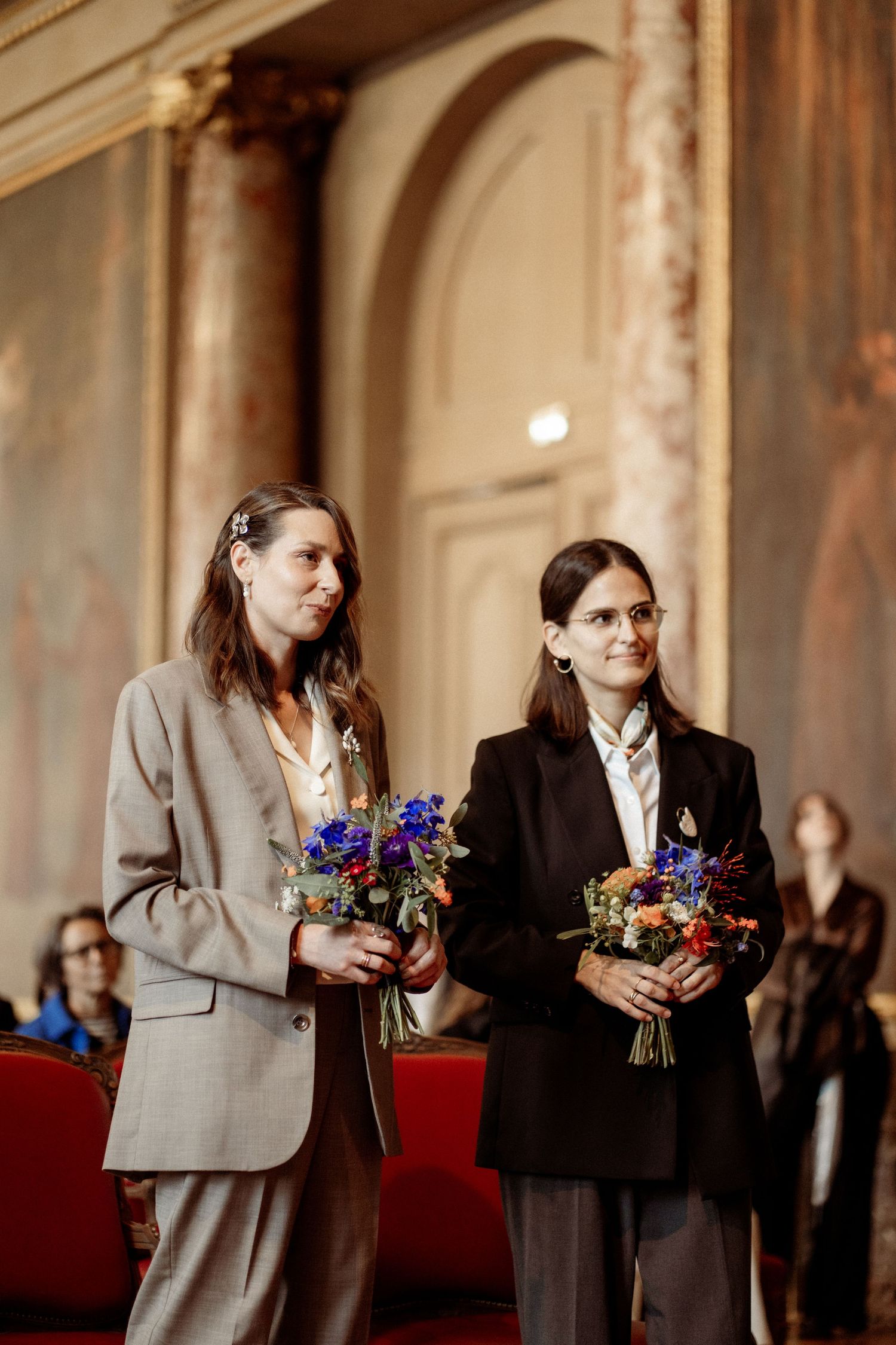 Two people in suits holding colorful flower bouquets stand in an ornate golden hall during a wedding ceremony.