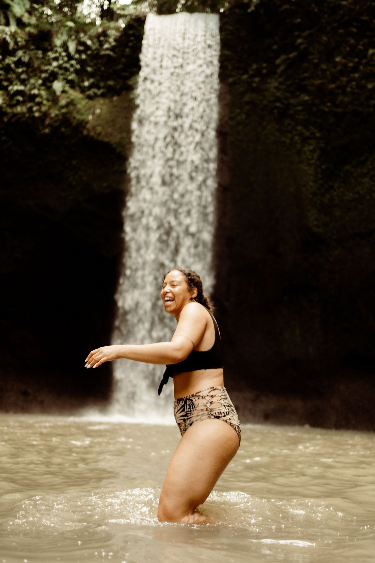A joyful woman in a black bikini top and animal print bottoms splashing in a pool beneath a lush tropical waterfall.