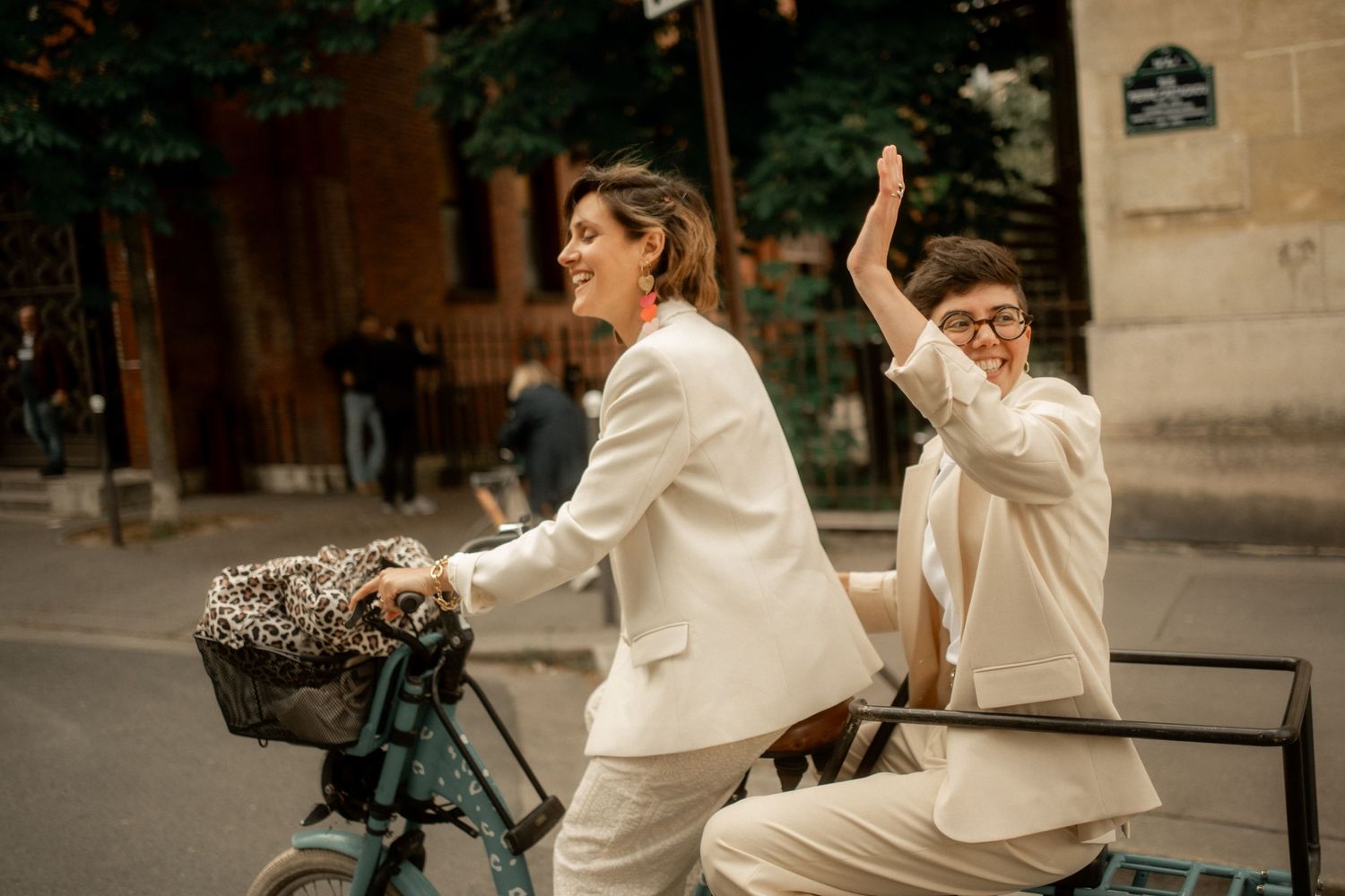 Two joyful women in cream suits ride a bicycle together on a Parisian street, laughing and high-fiving.