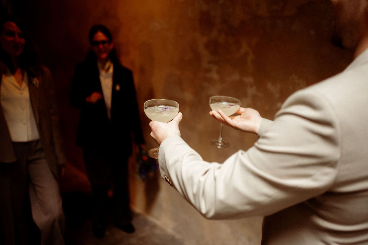Person holding two cocktail glasses in warm ambient light at an elegant wedding reception.