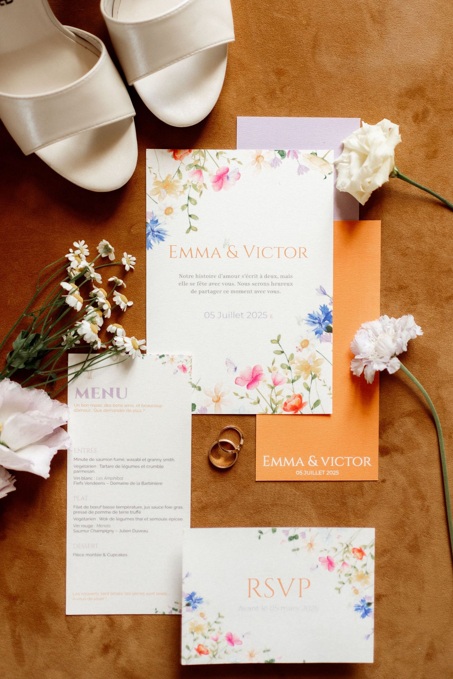 Wedding invitation suite with floral designs in coral and blue tones displayed on brown leather surface with rings and flowers.