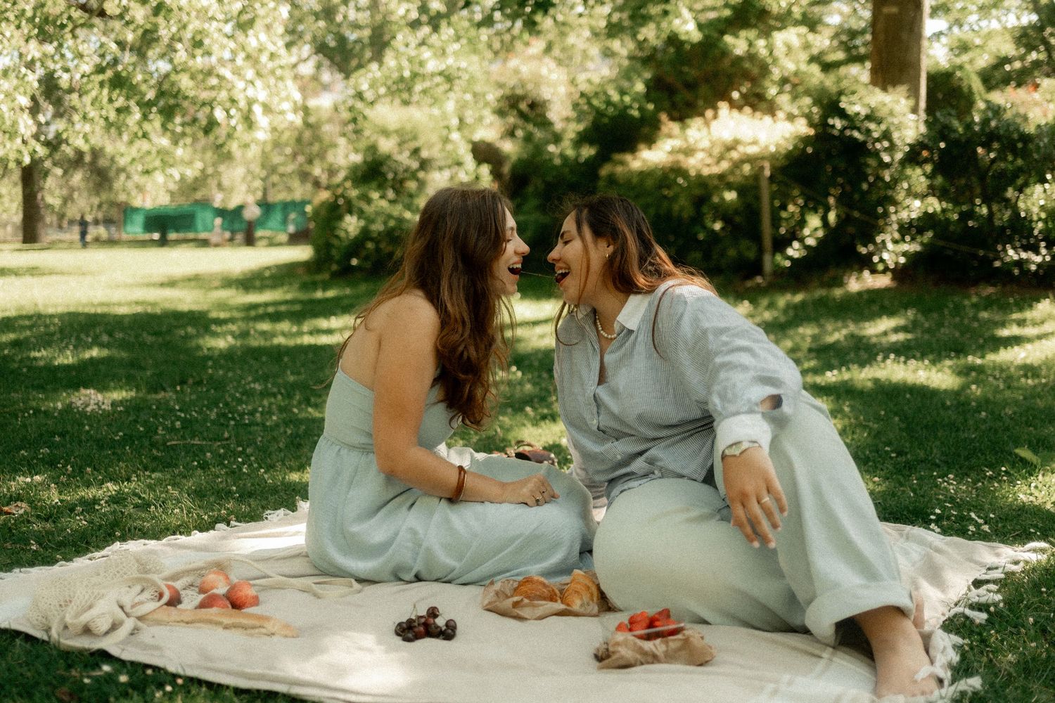 Two women laughing together on a picnic blanket in a lush green park on a sunny day.