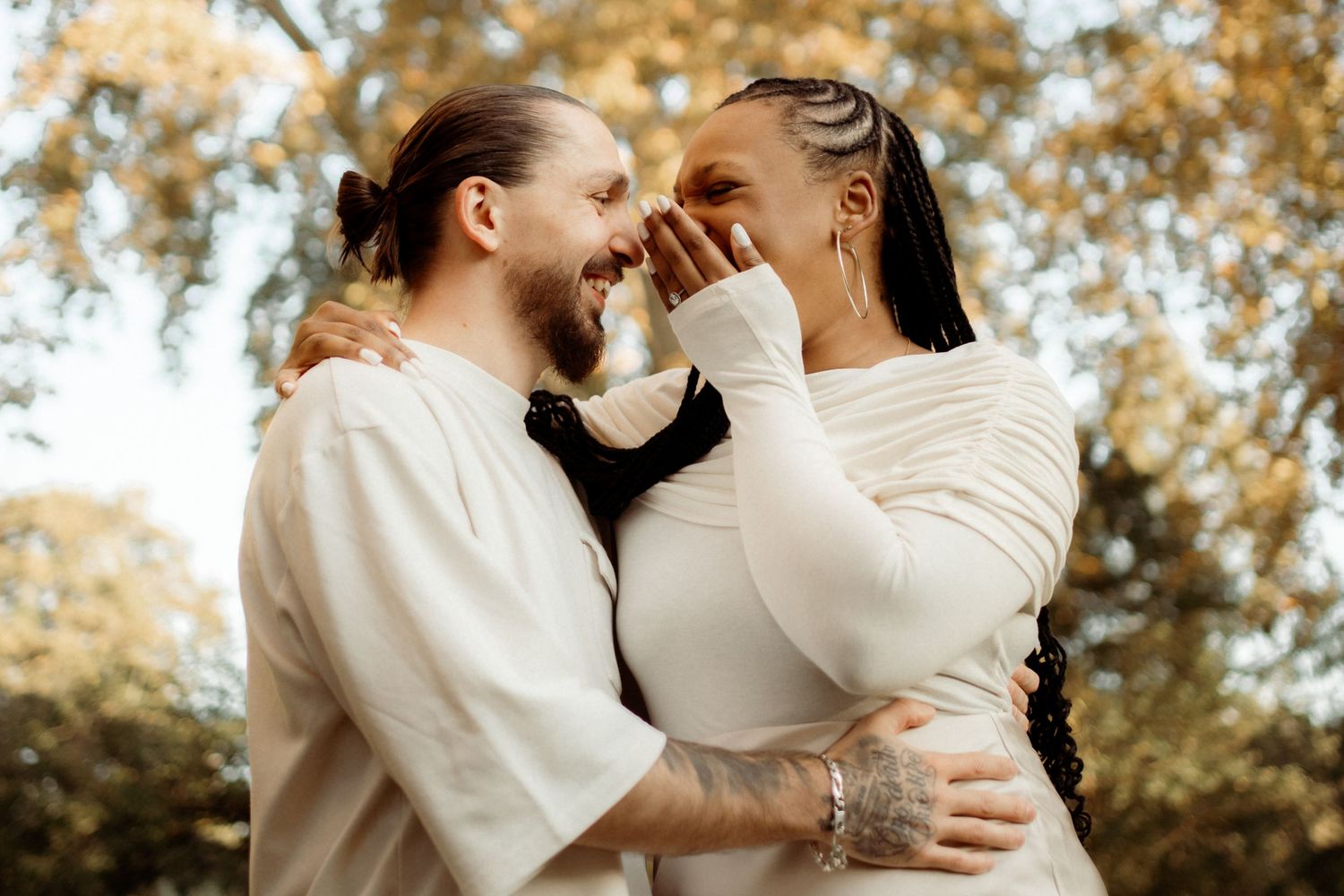 A couple embraces and smiles at each other outdoors, surrounded by autumn foliage, wearing matching cream outfits.