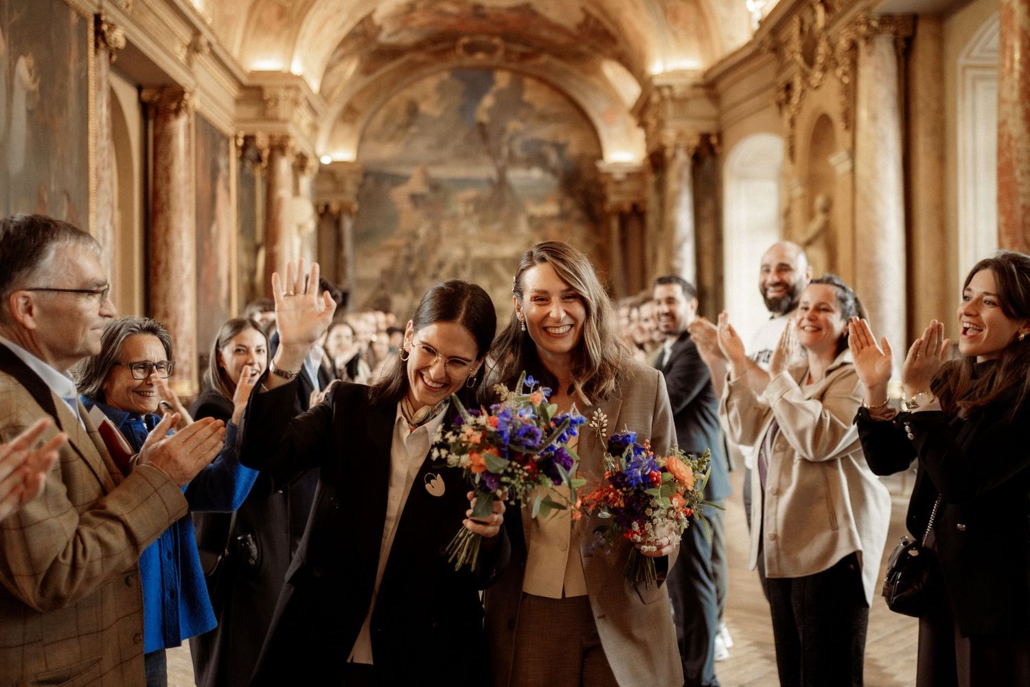 Two brides walk joyfully through ornate baroque hall, guests applauding, one holding colorful wildflower bouquet.