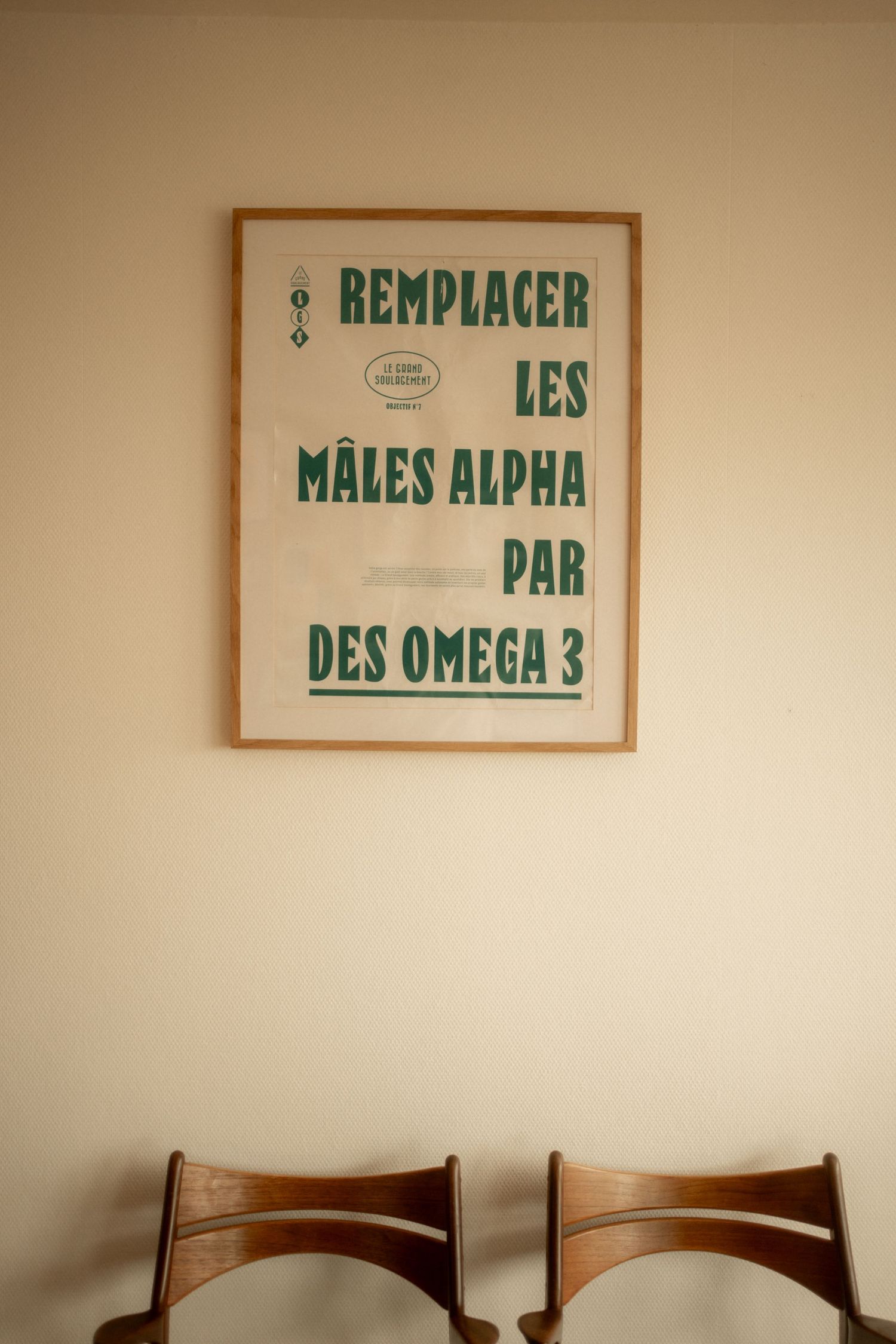 Framed French typography poster reading Remplacer Les Males Alpha Par Des Omega 3 hanging on a beige wall above two wooden chairs.