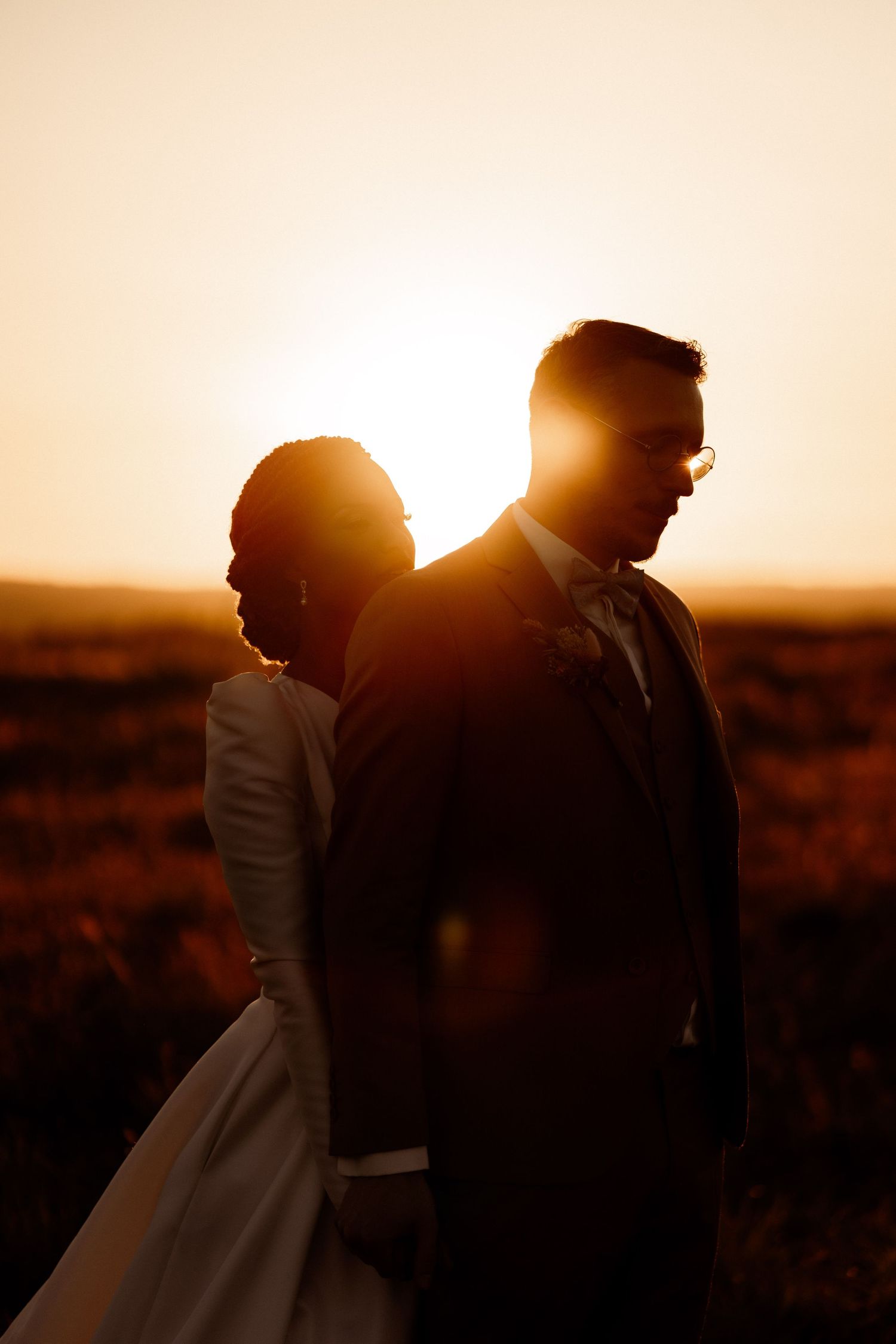 Silhouetted couple in wedding attire stand back-to-back against golden sunset over field.
