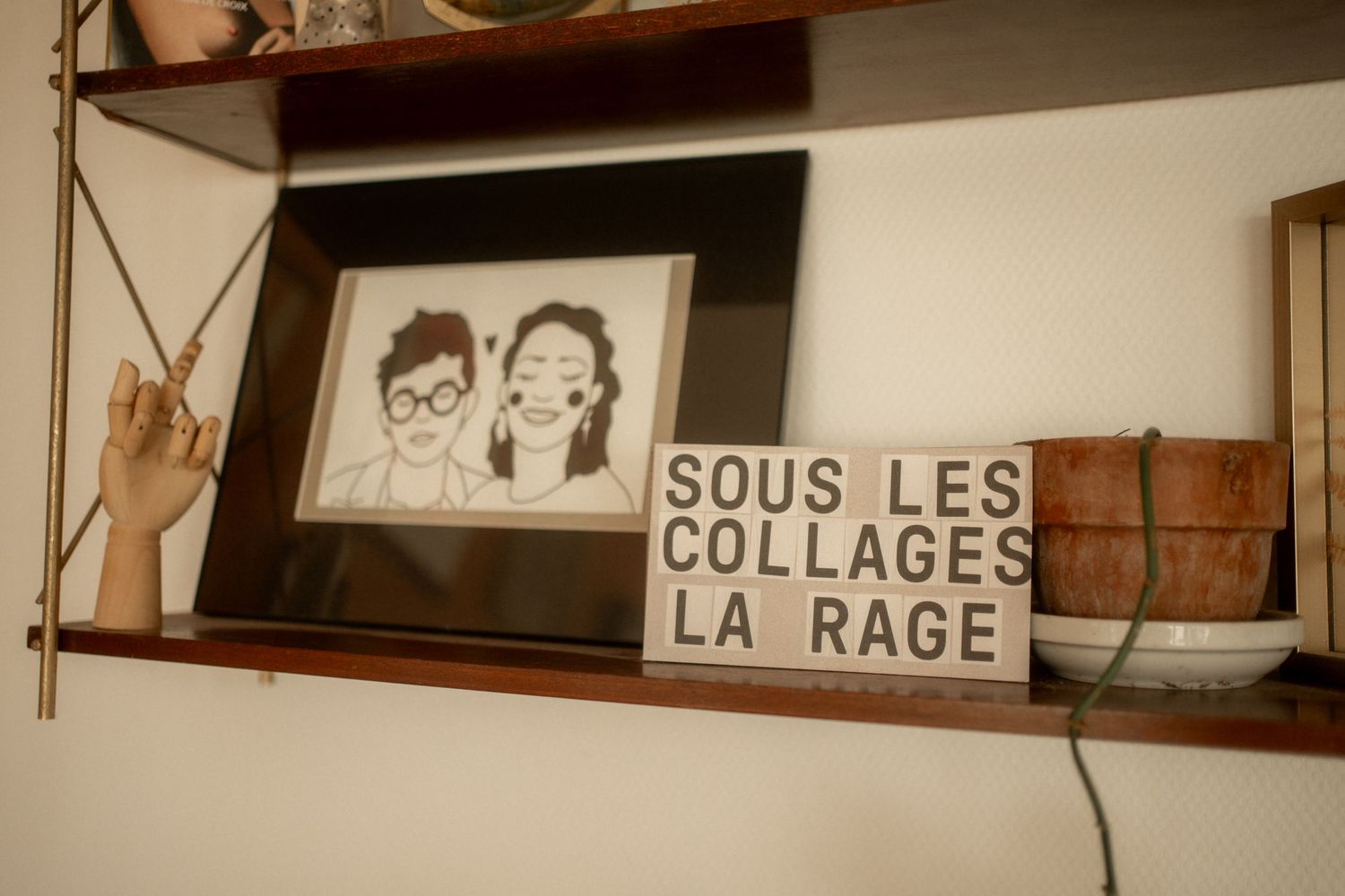 Wooden shelf displaying a framed portrait illustration and a sign reading Sous Les Collages La Rage.