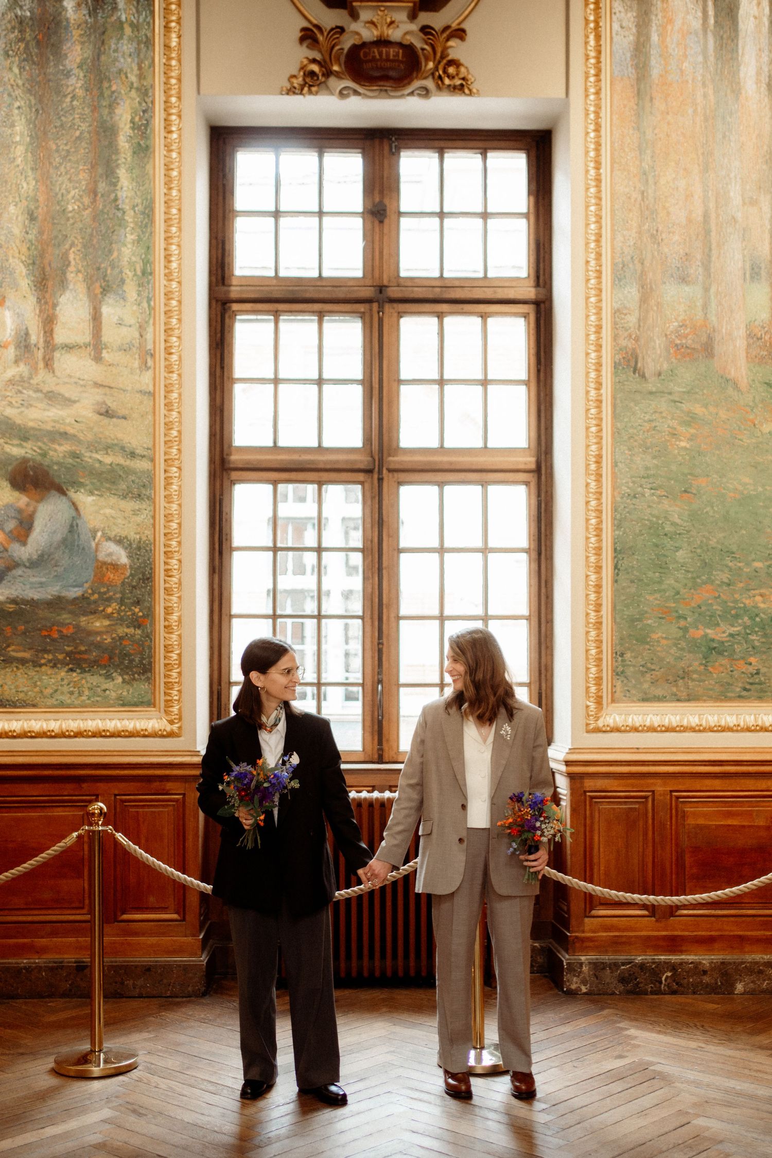 Two women holding flowers standing in elegant wood-paneled hall with large windows and paintings.