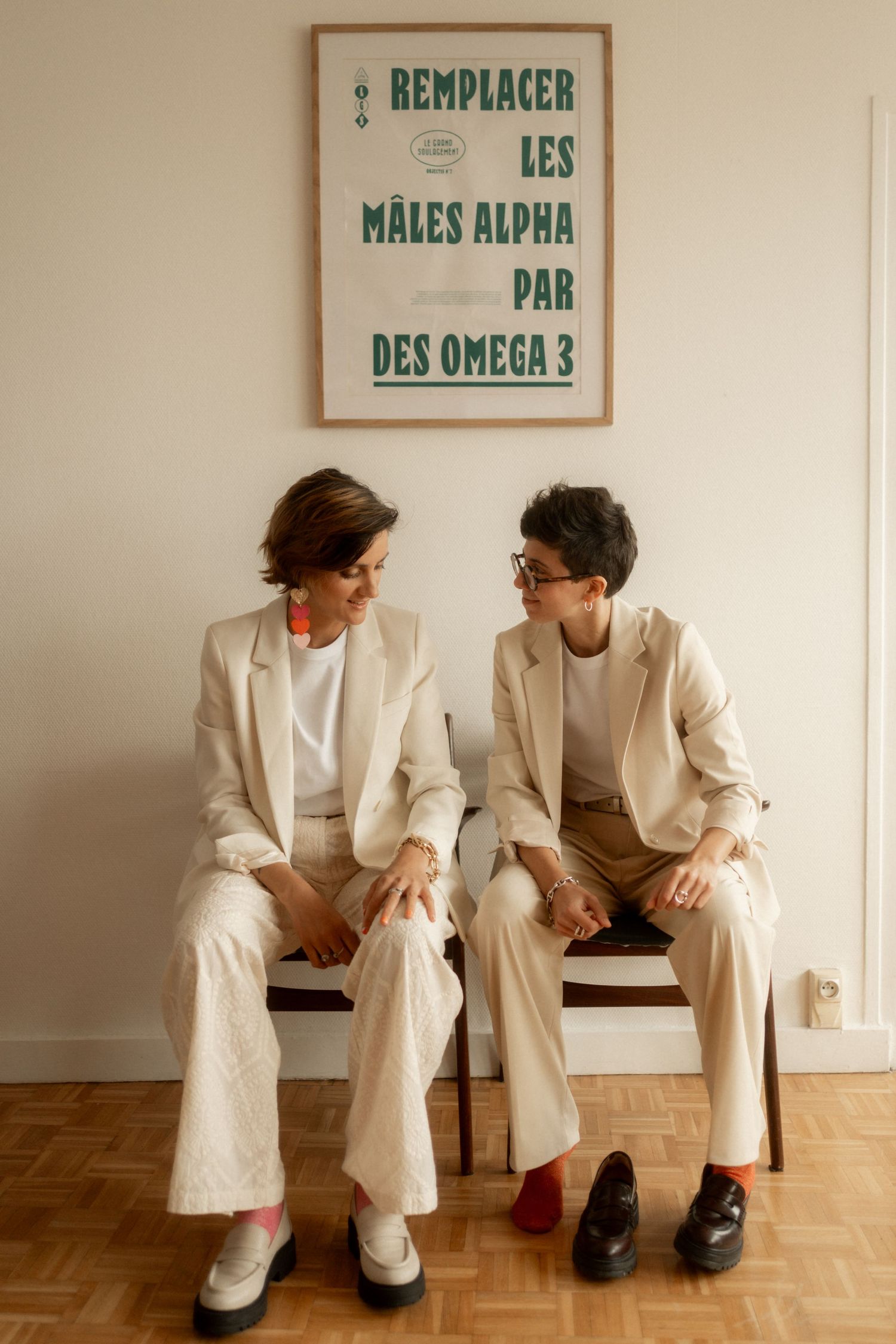 Two women in matching cream suits sit on chairs, smiling at each other beneath a French feminist poster.