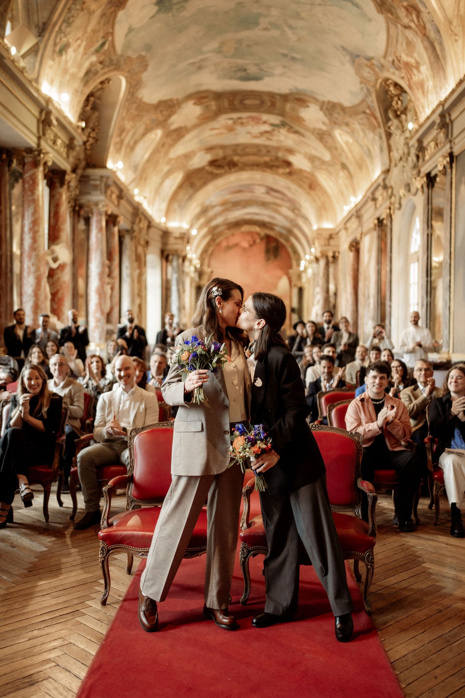 Two brides share a kiss at their wedding ceremony in an ornate baroque hall with frescoed ceilings.