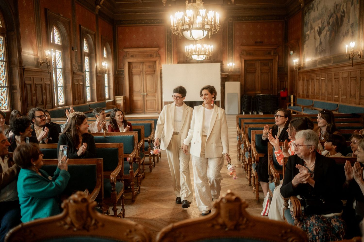 Two brides in white suits walk hand in hand down the aisle in an elegant wood-paneled wedding hall.
