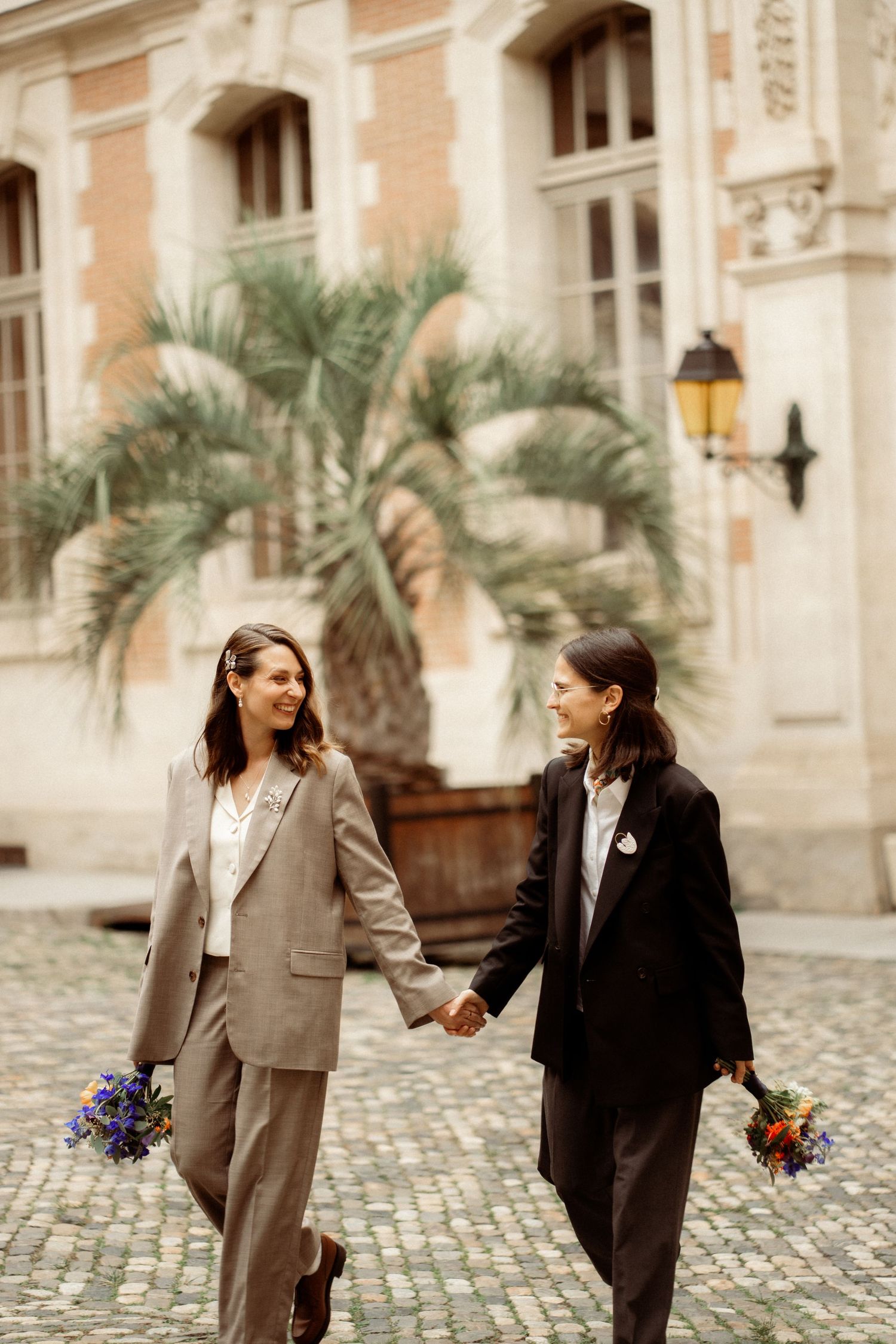 Two brides in suits holding hands and bouquets walking on cobblestone courtyard with palm tree.