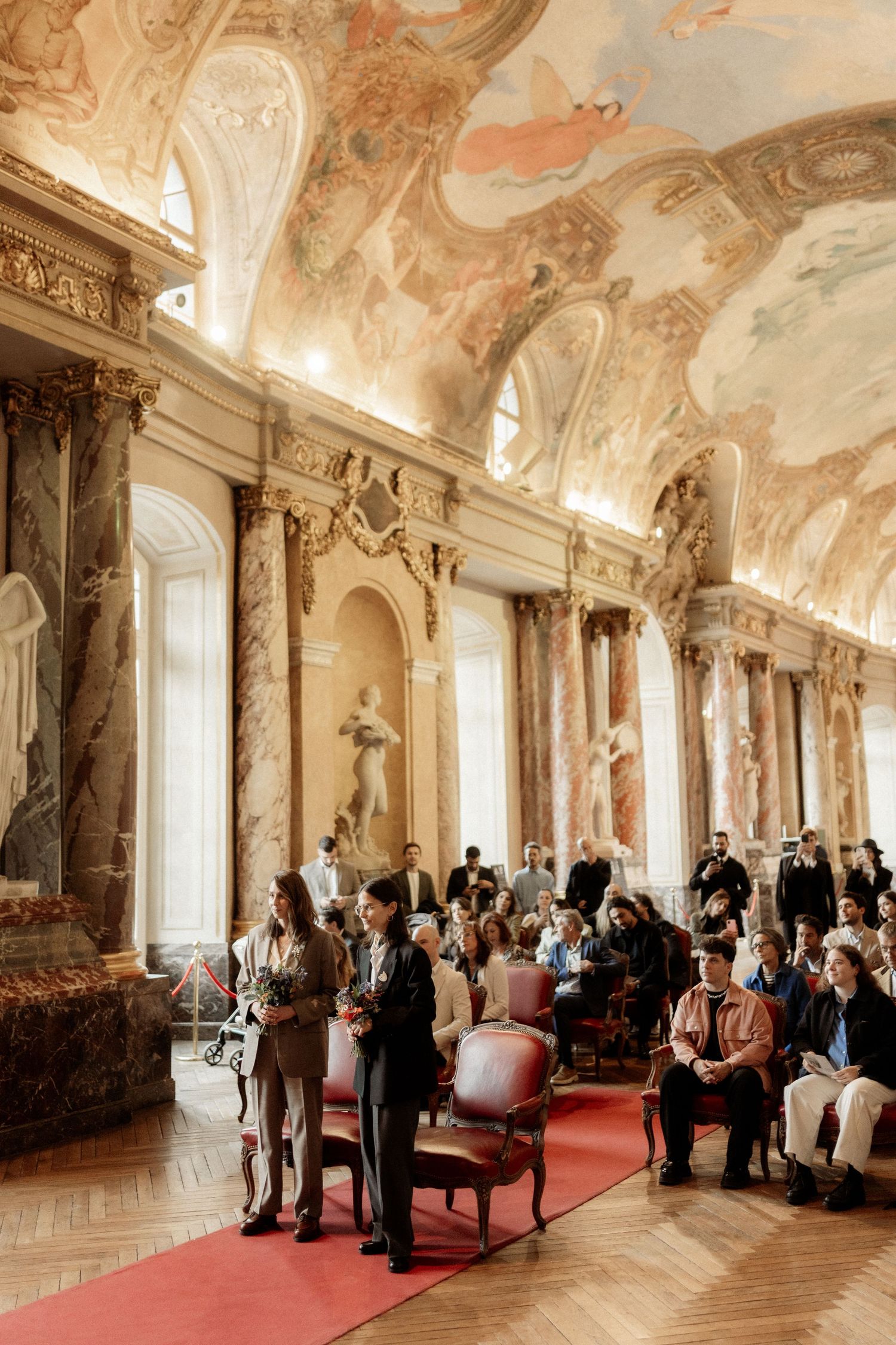 Elegant wedding ceremony in a grand baroque hall with marble columns and ornate frescoed ceiling.