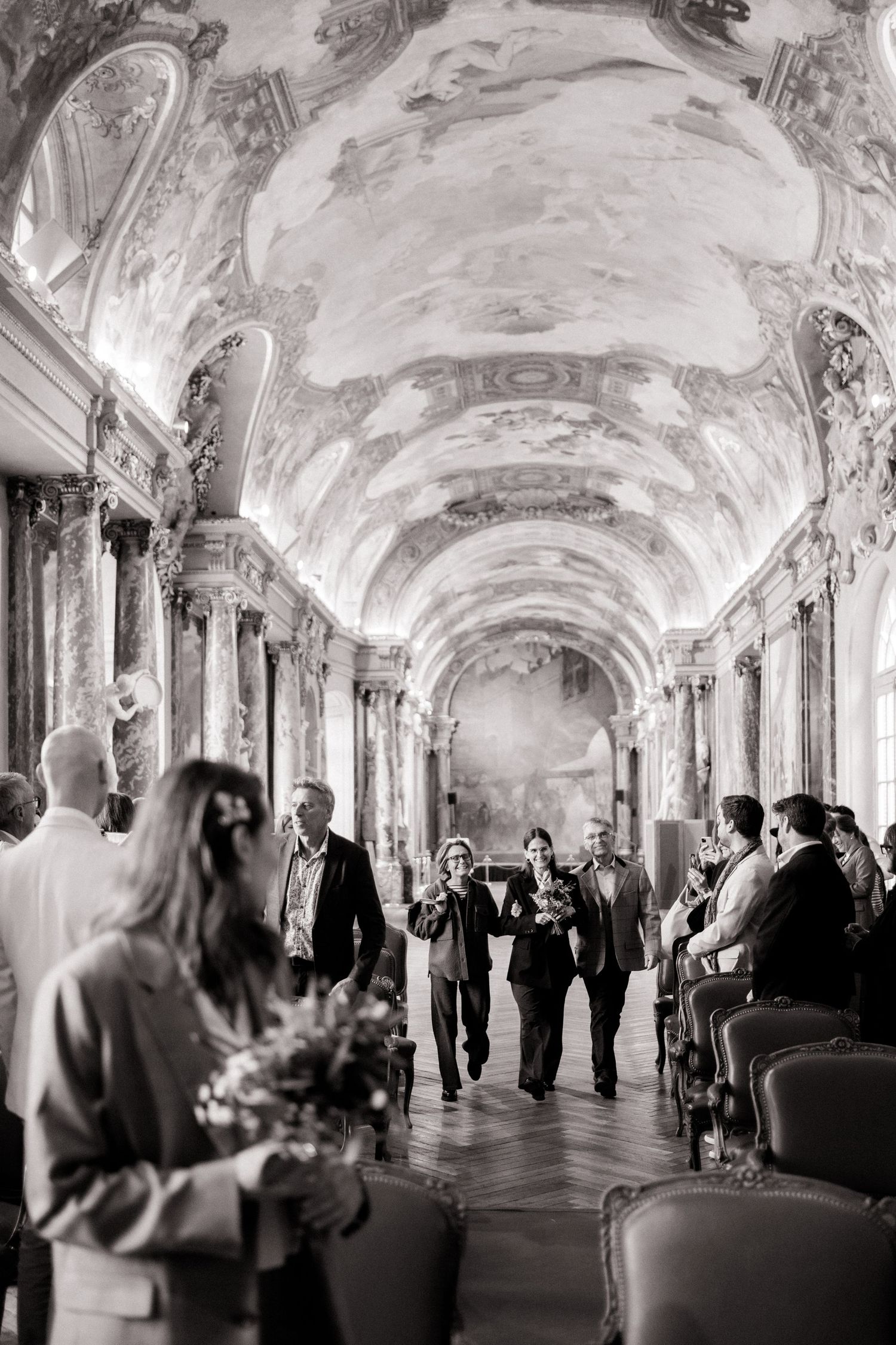 Black and white photo of wedding guests walking down ornate baroque hall with frescoed ceiling.