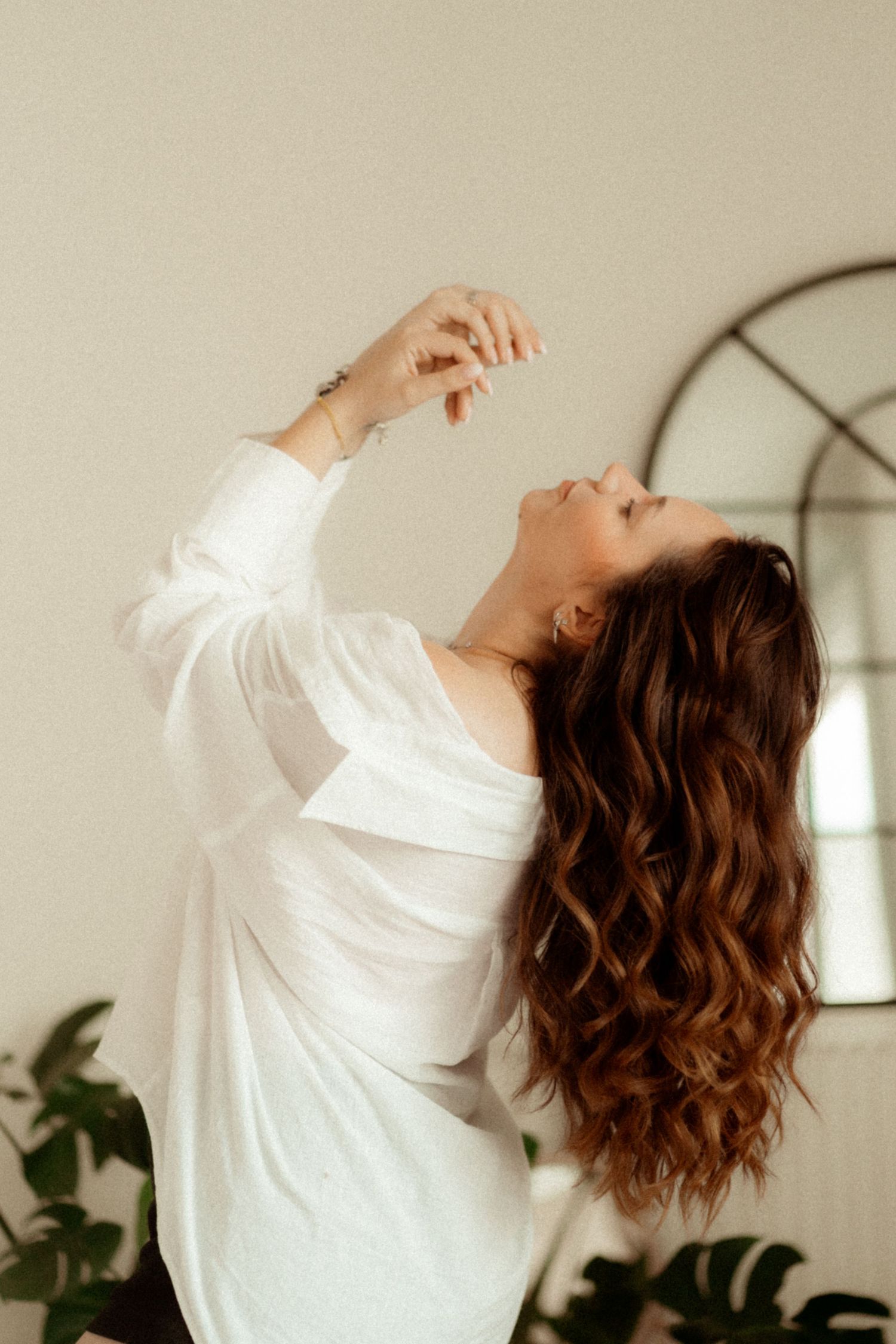 A person in a white shirt with long wavy brown hair poses gracefully against a beige wall with indoor plants and mirror.