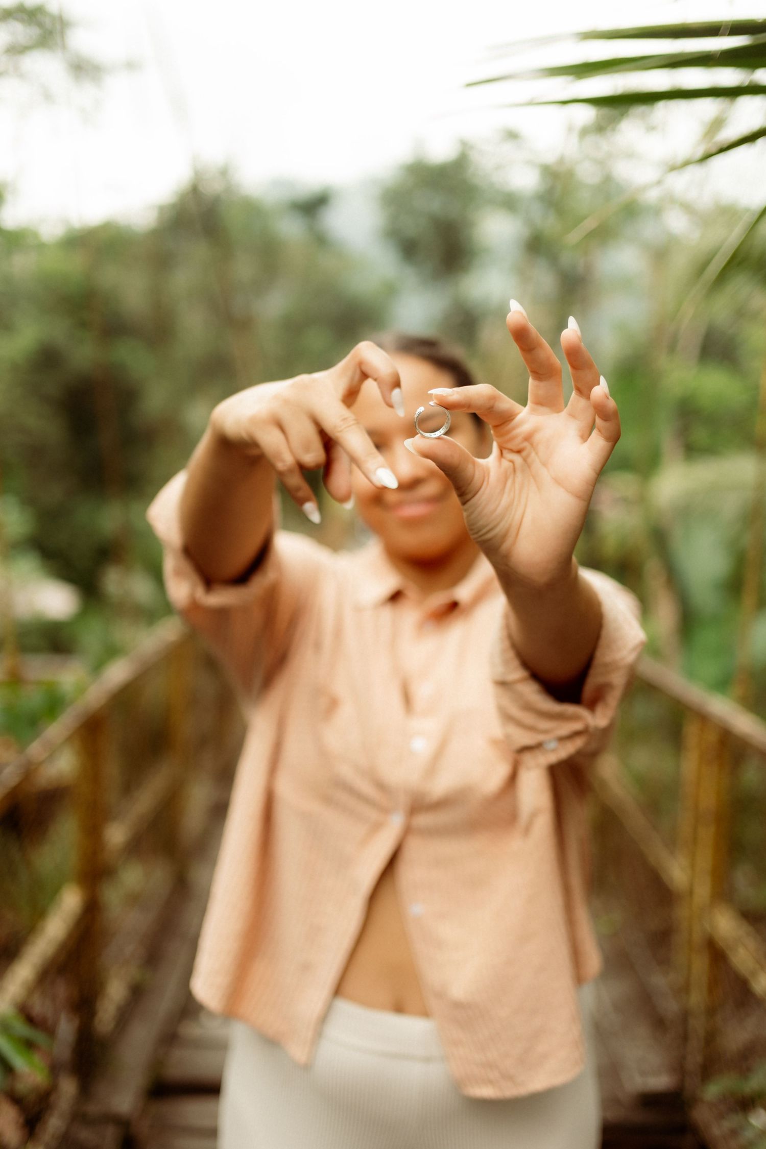 Woman in peach shirt poses with hands outstretched toward camera in lush tropical garden setting.
