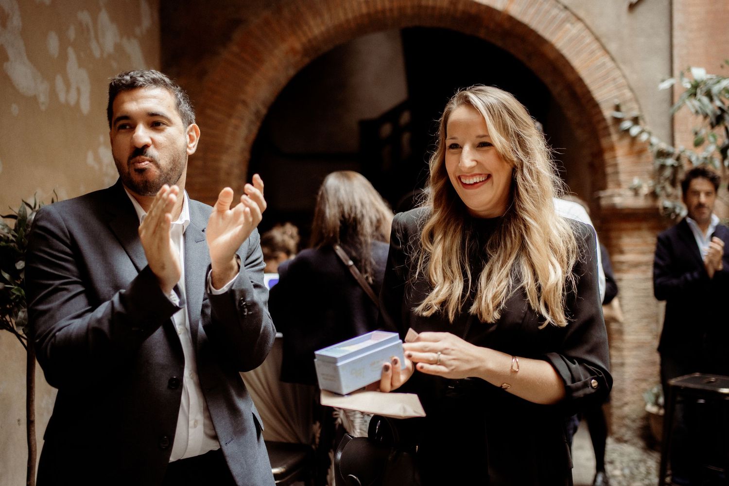 Man and woman smiling at an indoor event, man clapping in a black blazer, woman holding a small box.