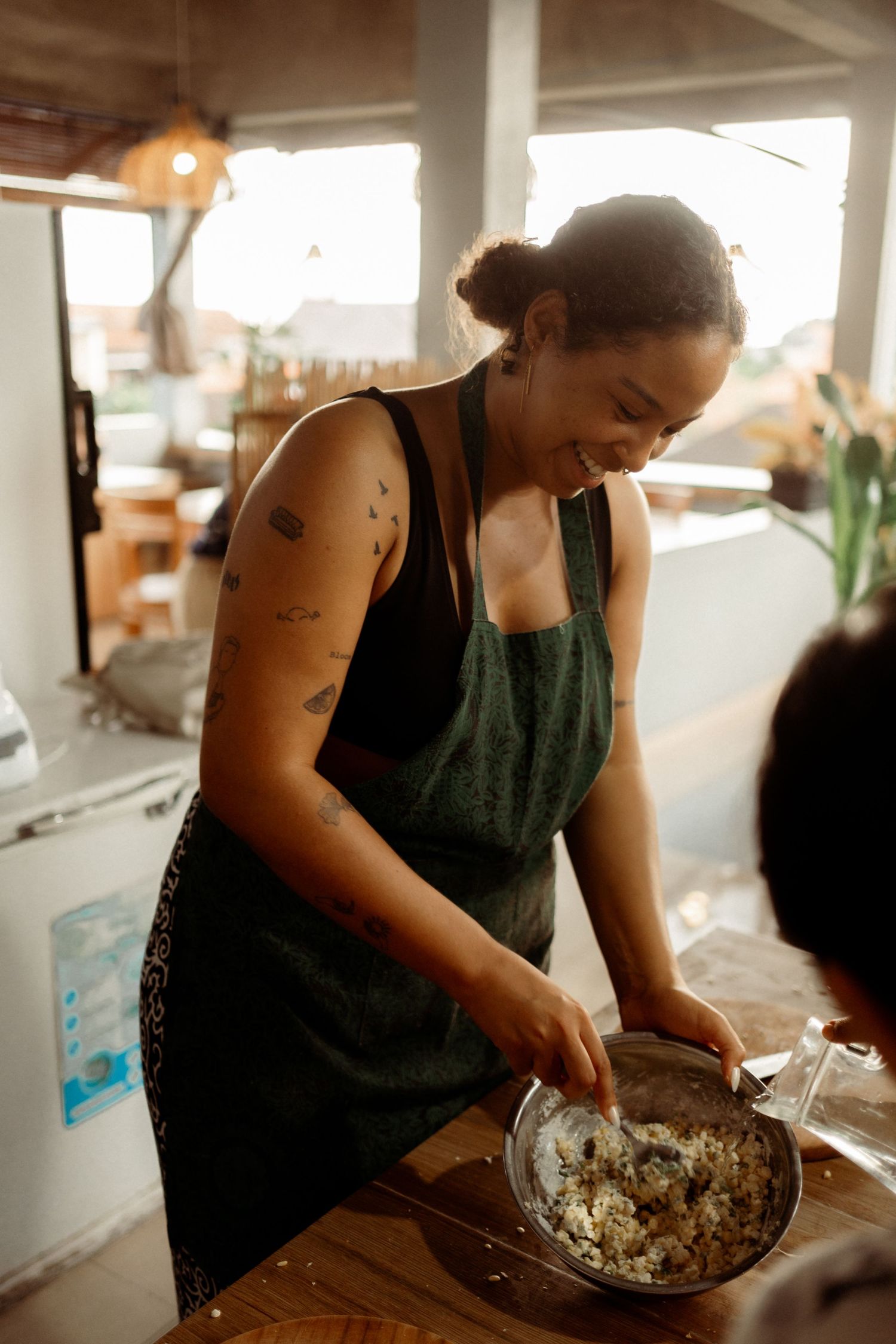 A smiling woman wearing a green apron mixes ingredients in a bowl at a wooden kitchen counter.