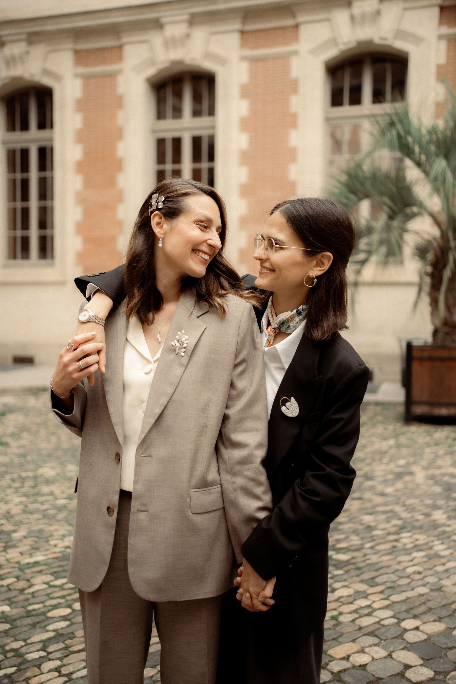 Two women in tailored suits hold hands and smile at each other in a cobblestone European courtyard.