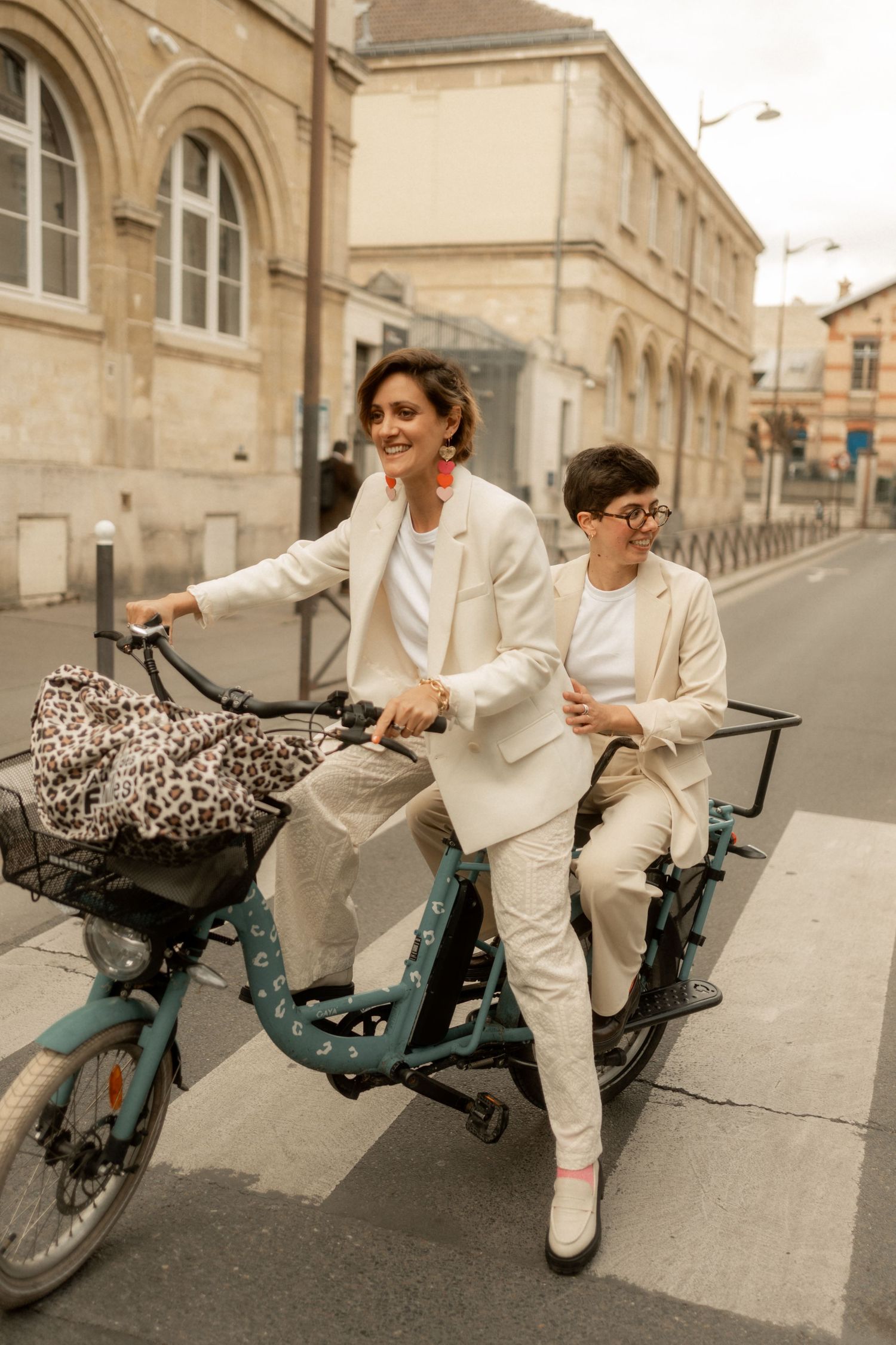 Two women in matching cream blazers riding a cargo bike together on a Parisian street.