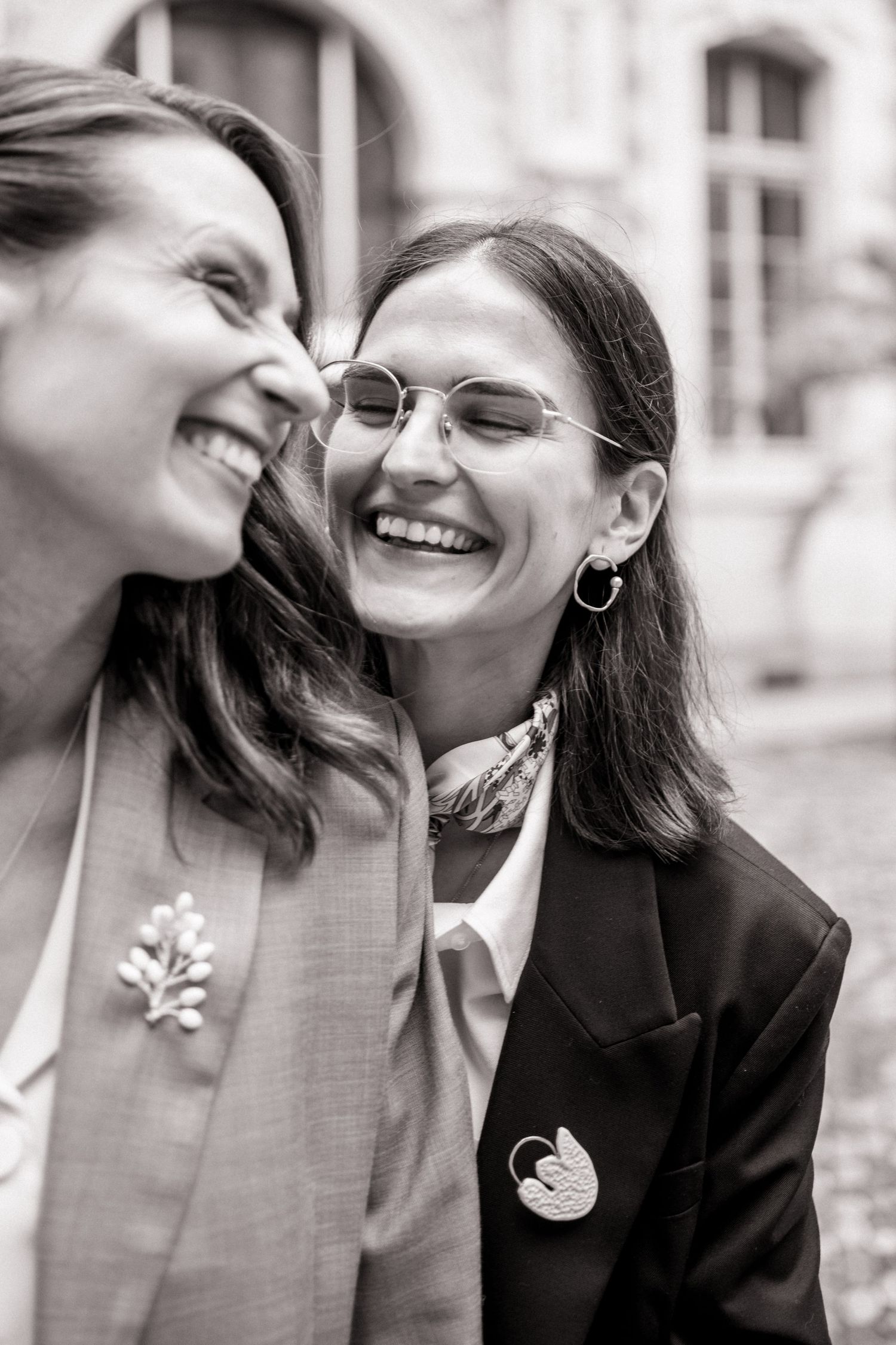 Black and white close-up of two laughing women wearing brooches celebrating at an outdoor wedding.