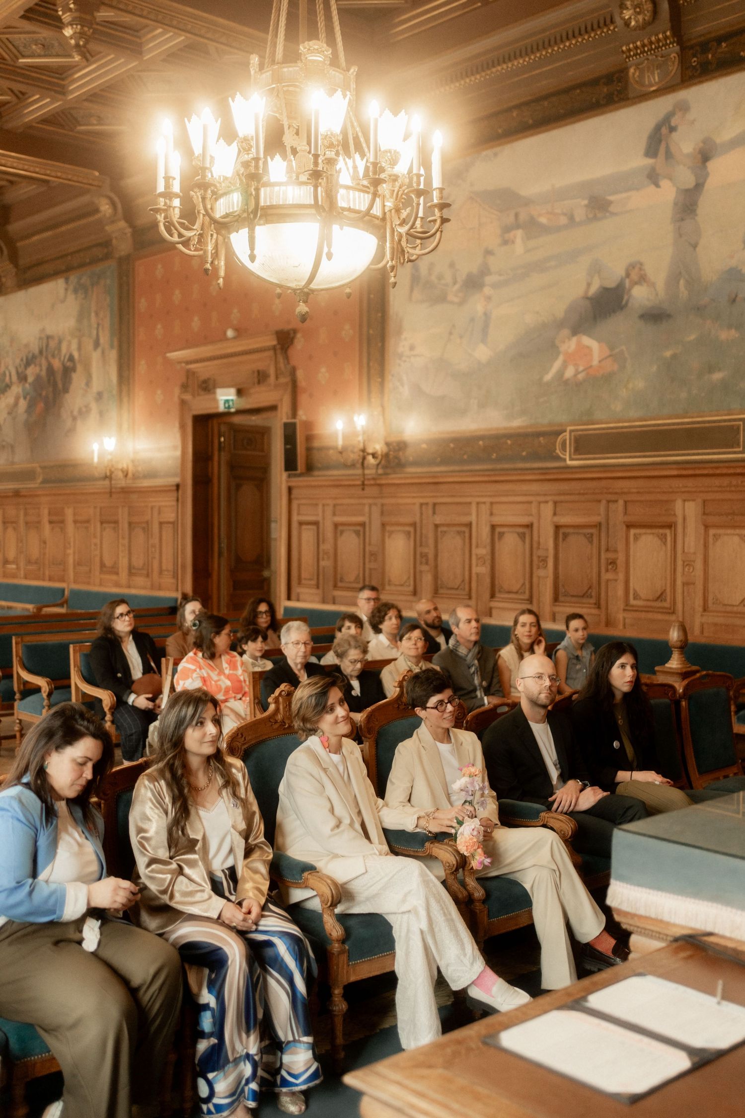 Guests seated in an ornate wood-paneled ceremony room with chandelier and murals during a wedding.