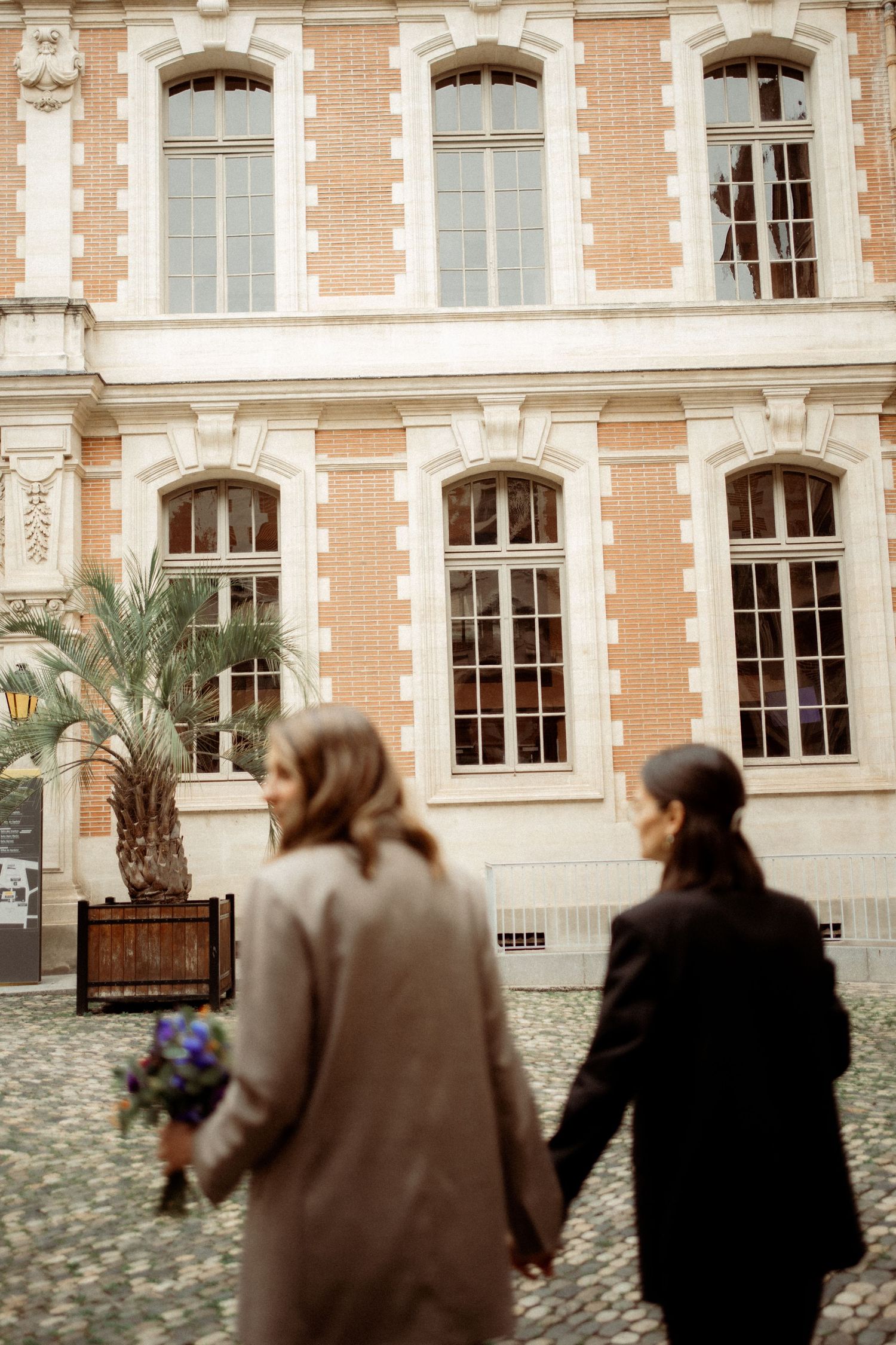 Two women holding hands walking toward an elegant brick building on a cobblestone street.